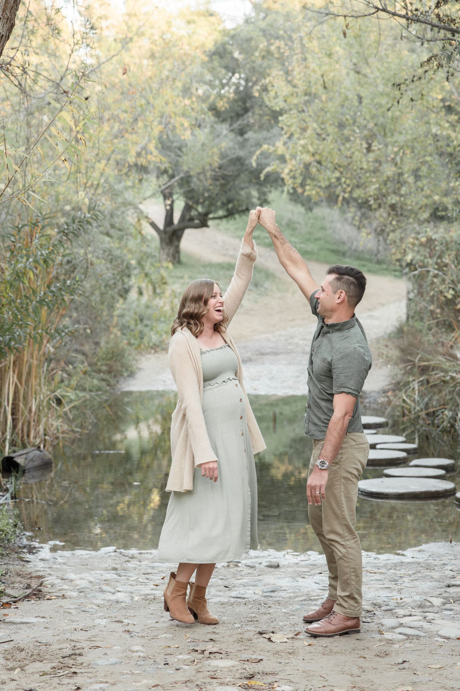 Pregnant woman in sage dress laughing as her partner twirls her near a tranquil creek surrounded by autumn trees.