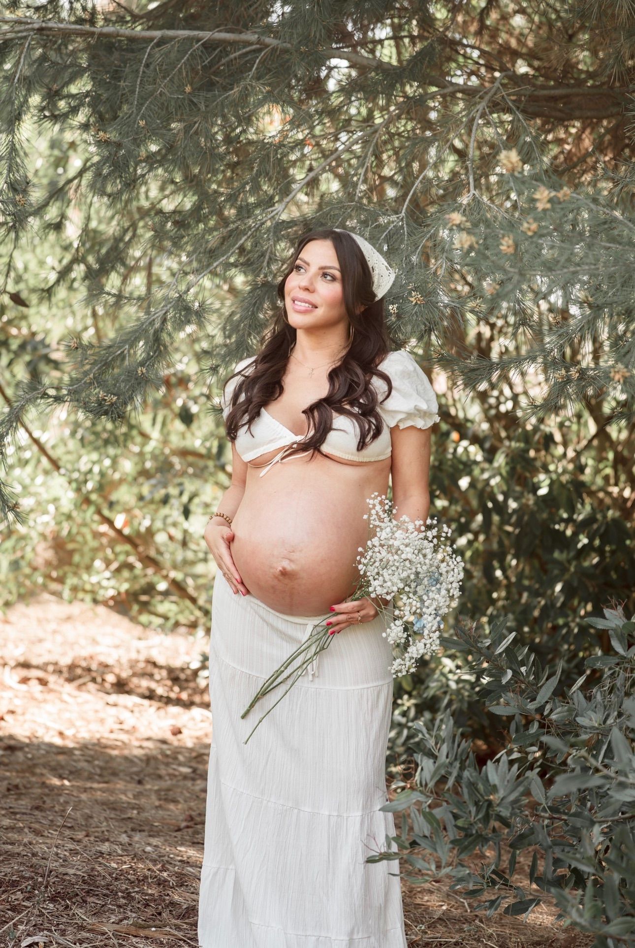 Pregnant woman in white crop top and maxi skirt holding baby's breath flowers outdoors among green trees.