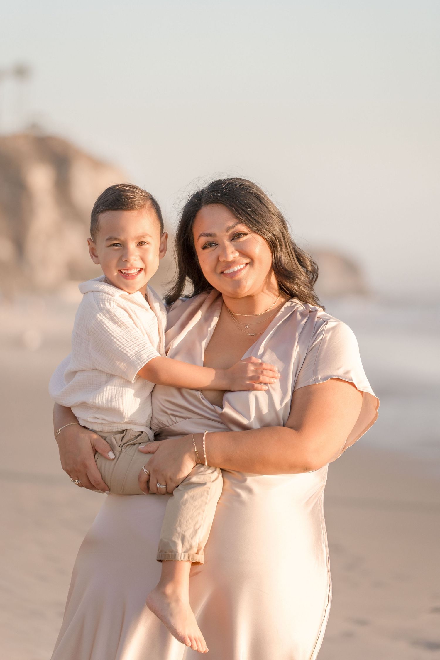 Smiling mother holding her young son on a sunny beach, both looking at the camera happily.