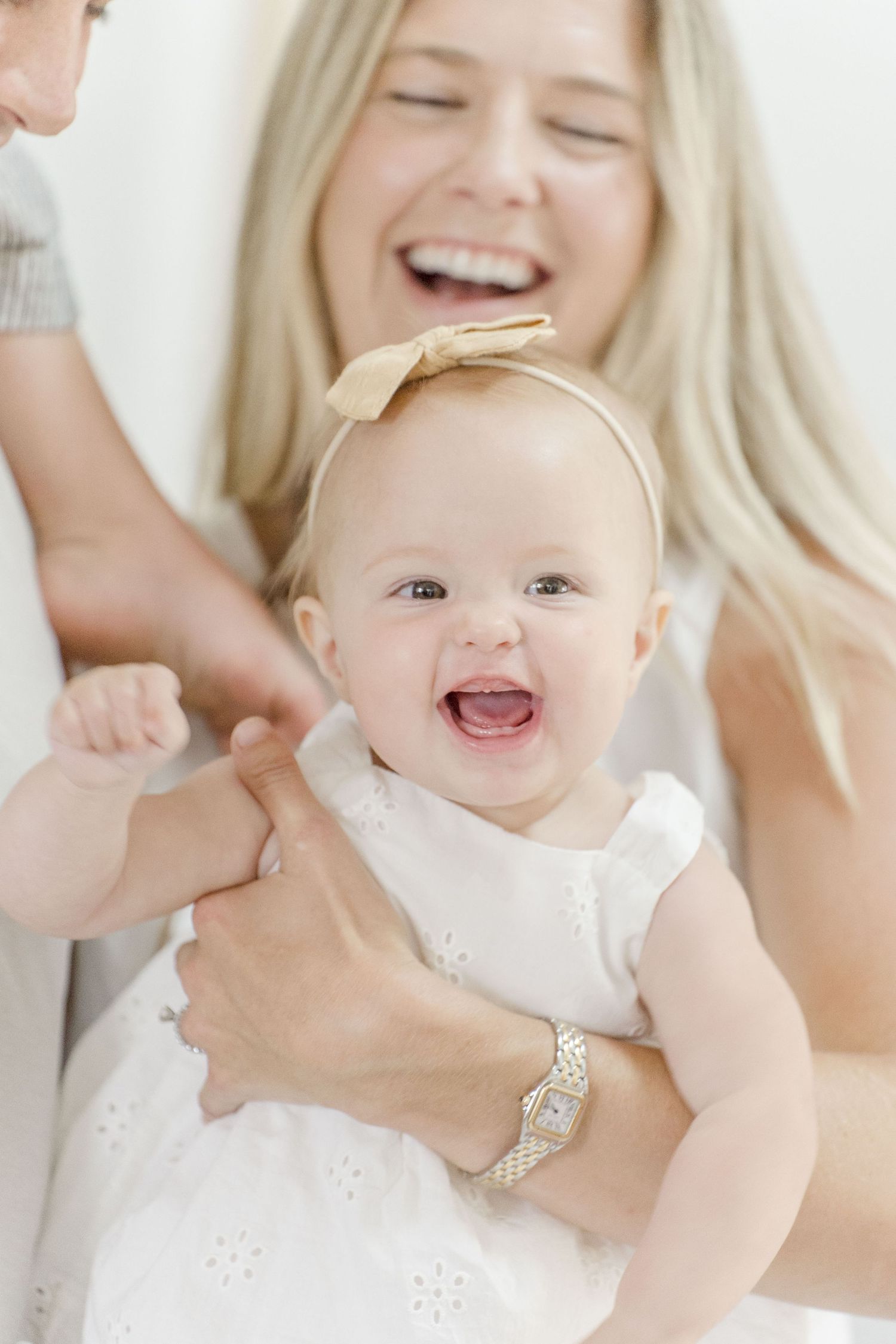 Laughing blonde mother holding a joyful baby girl in white dress with a gold bow headband.