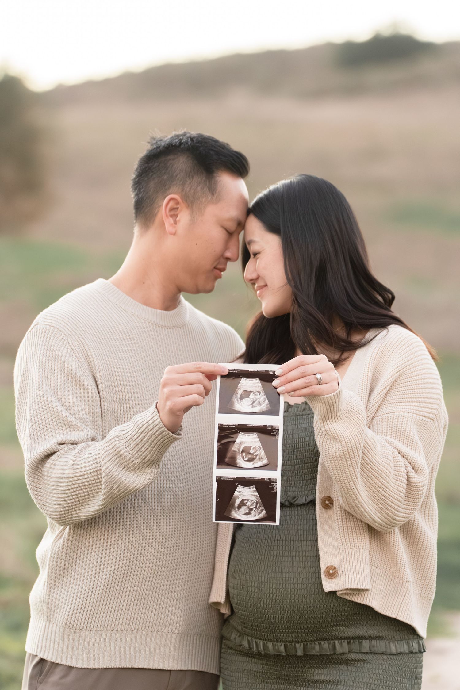 Couple touching foreheads while holding ultrasound photos outdoors in a golden field during maternity announcement session.