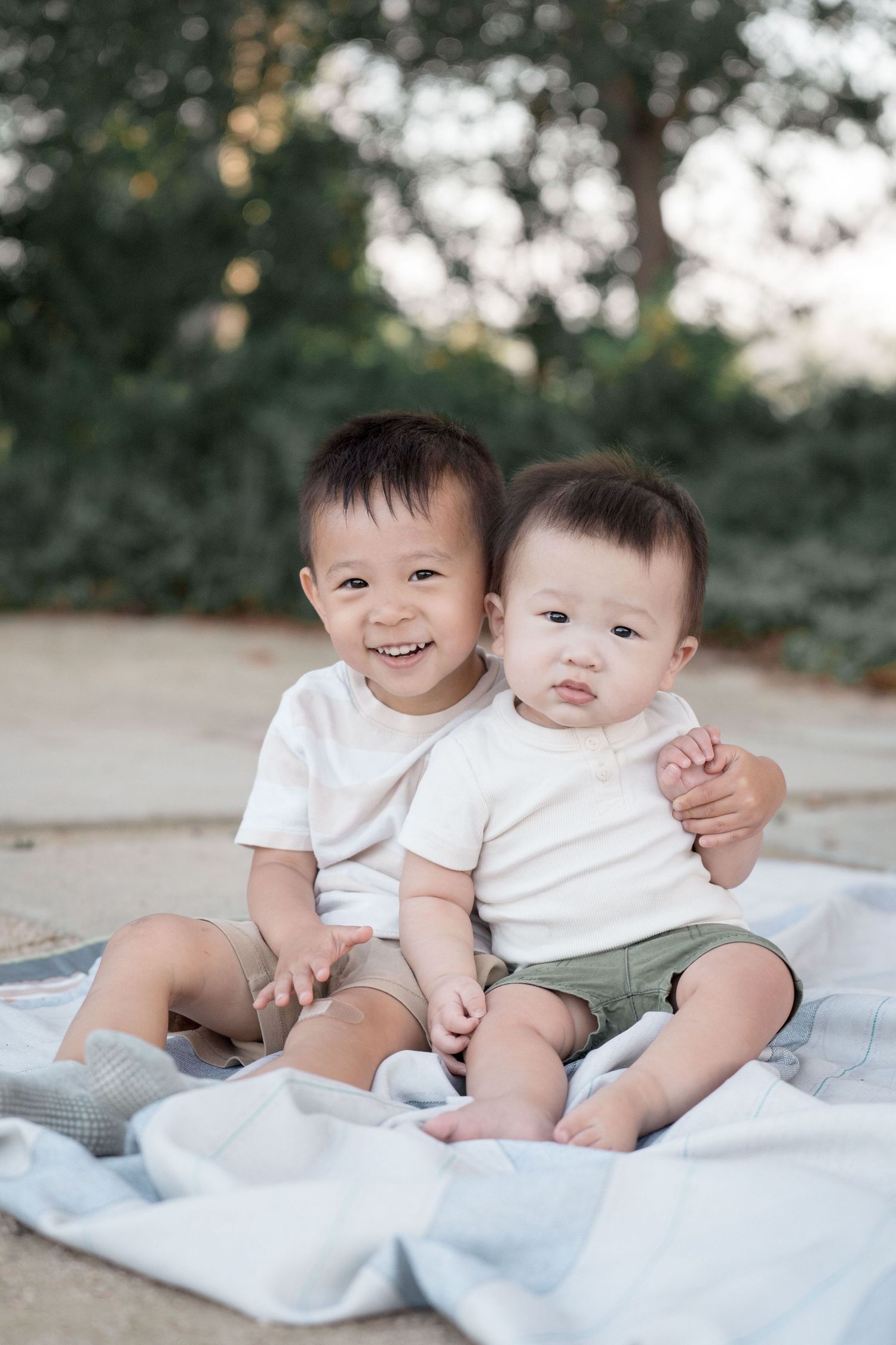 Two smiling Asian toddler brothers in white shirts sitting together on a blanket outdoors.