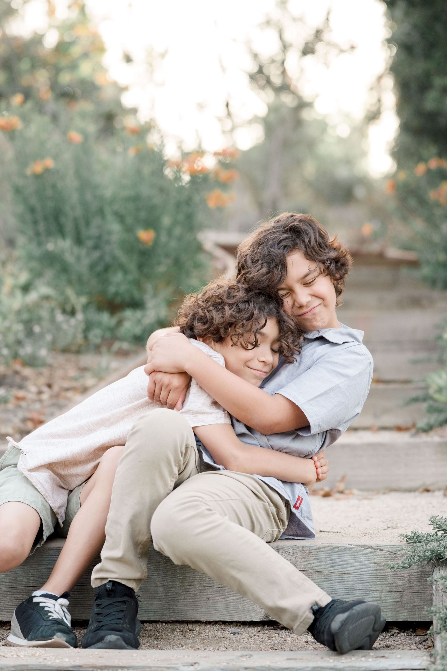 Two curly-haired brothers sharing a warm hug while sitting outdoors in a garden.