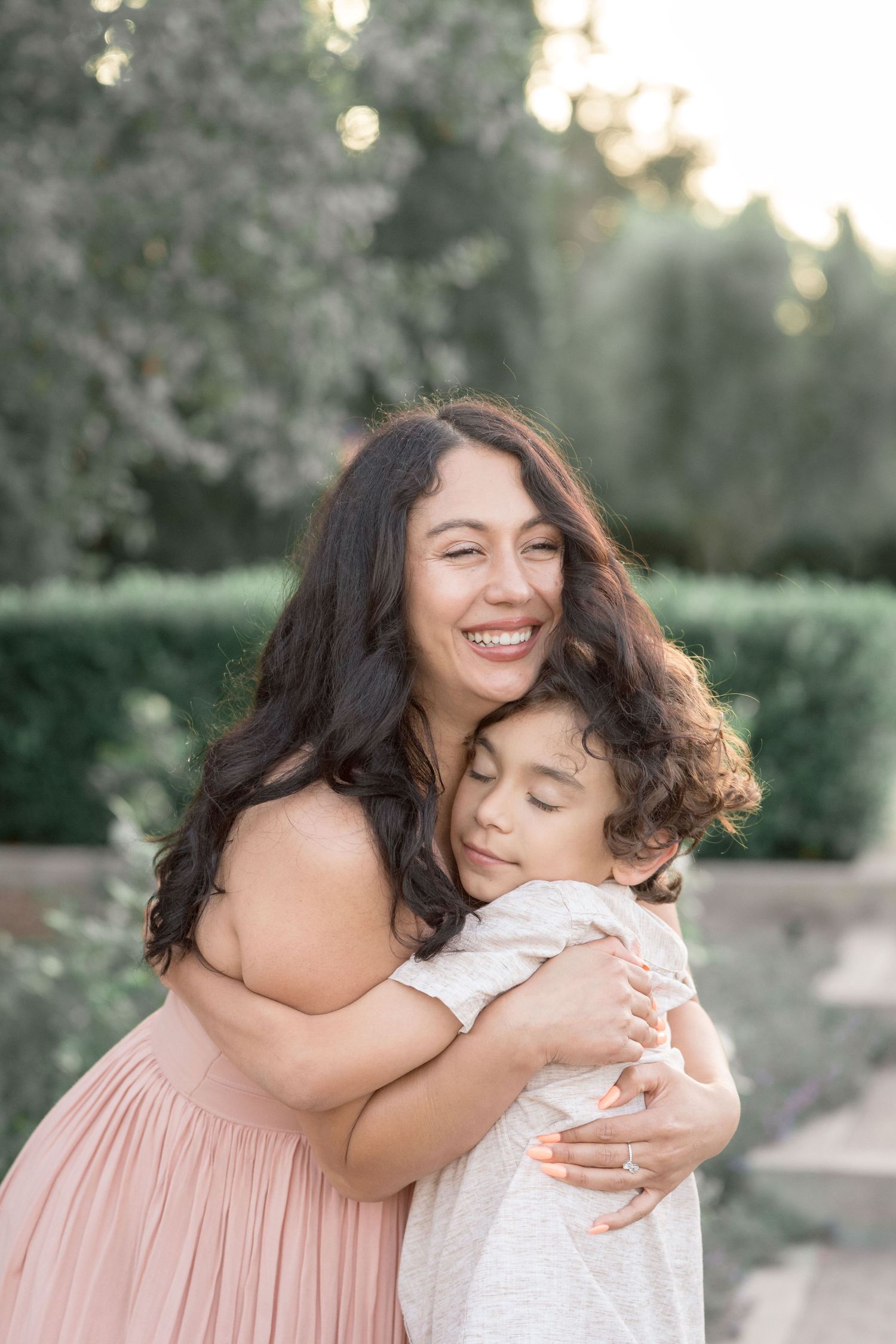 A joyful mother in a pink dress hugs her young child in a sunlit garden setting.