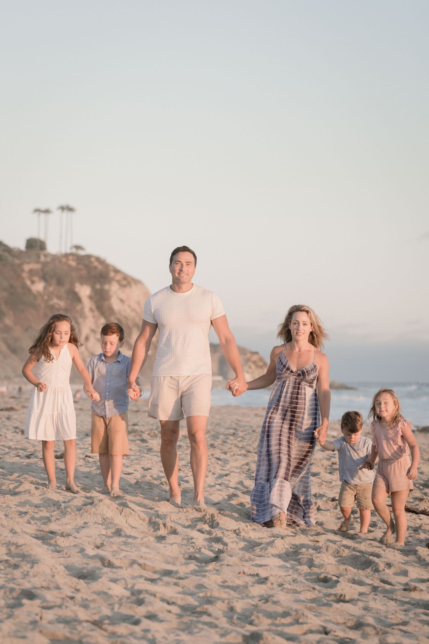 Happy family of six walking on a sandy beach at golden hour, parents holding hands with children.