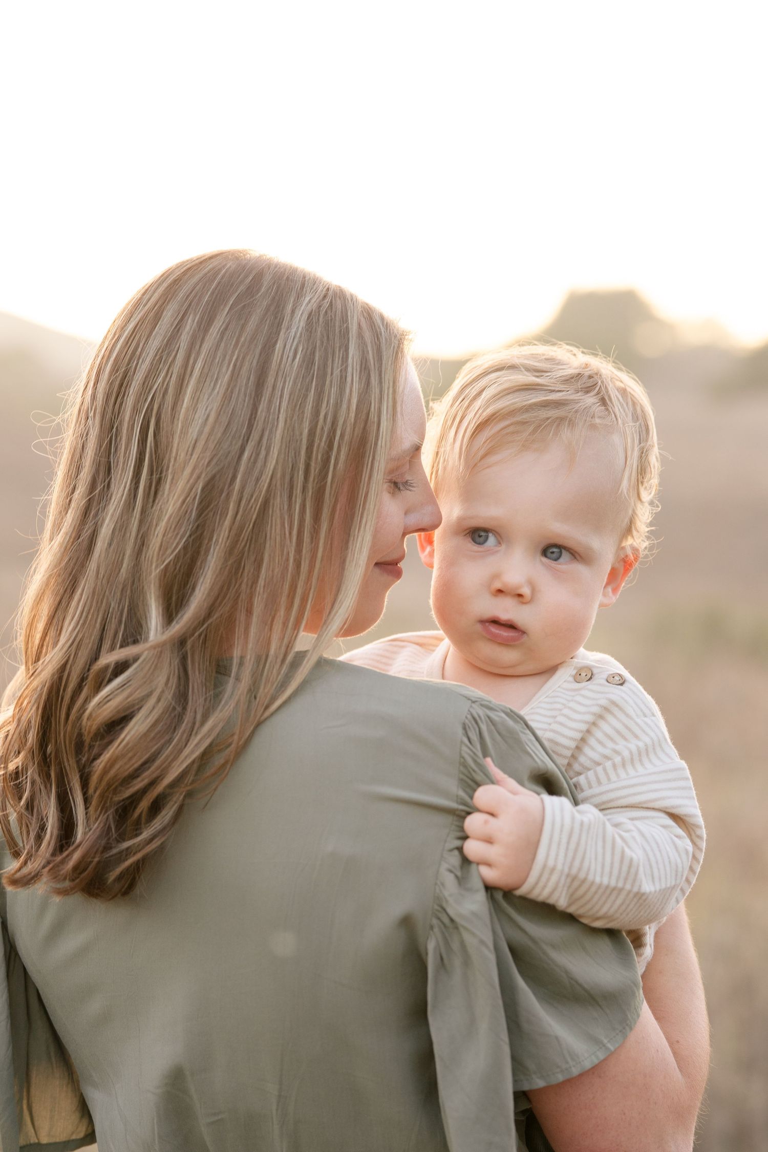 Blonde mother holds her wide-eyed baby boy against her shoulder in a sun-drenched open field.