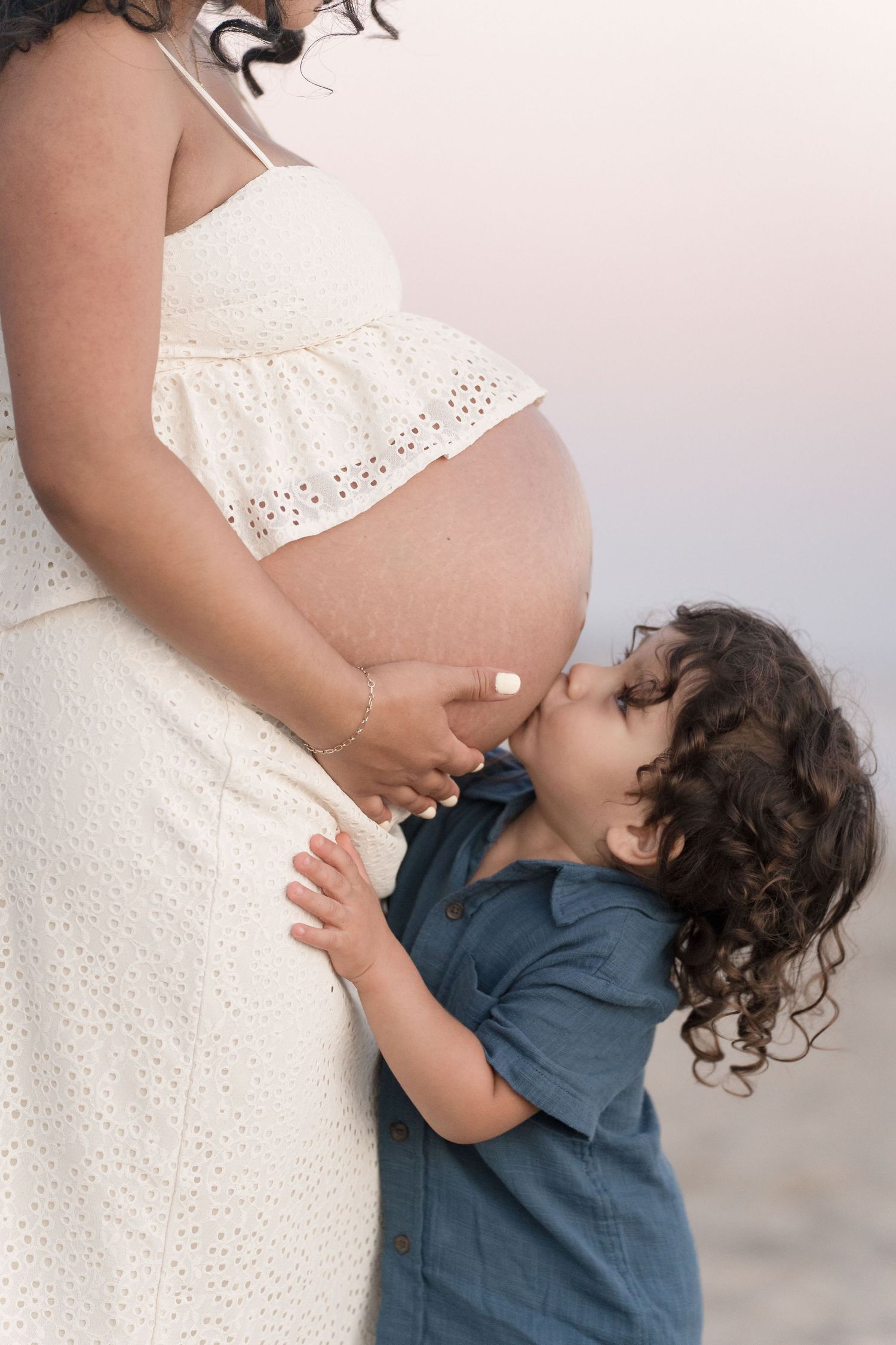 A curly-haired toddler sweetly kisses his pregnant mother\u2019s bare belly on the beach.