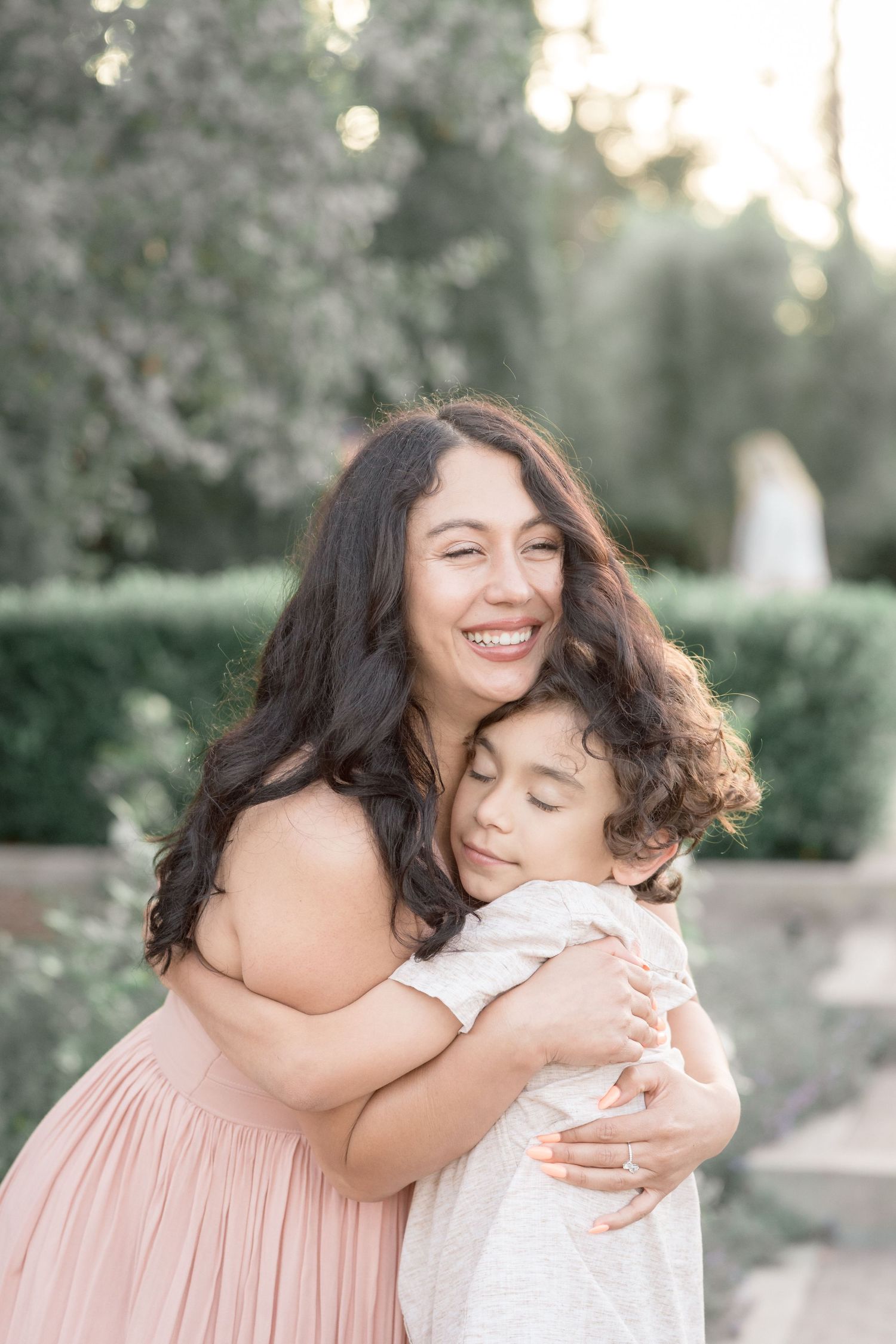 Joyful mother in pink dress hugging her young curly-haired child in a lush garden setting.