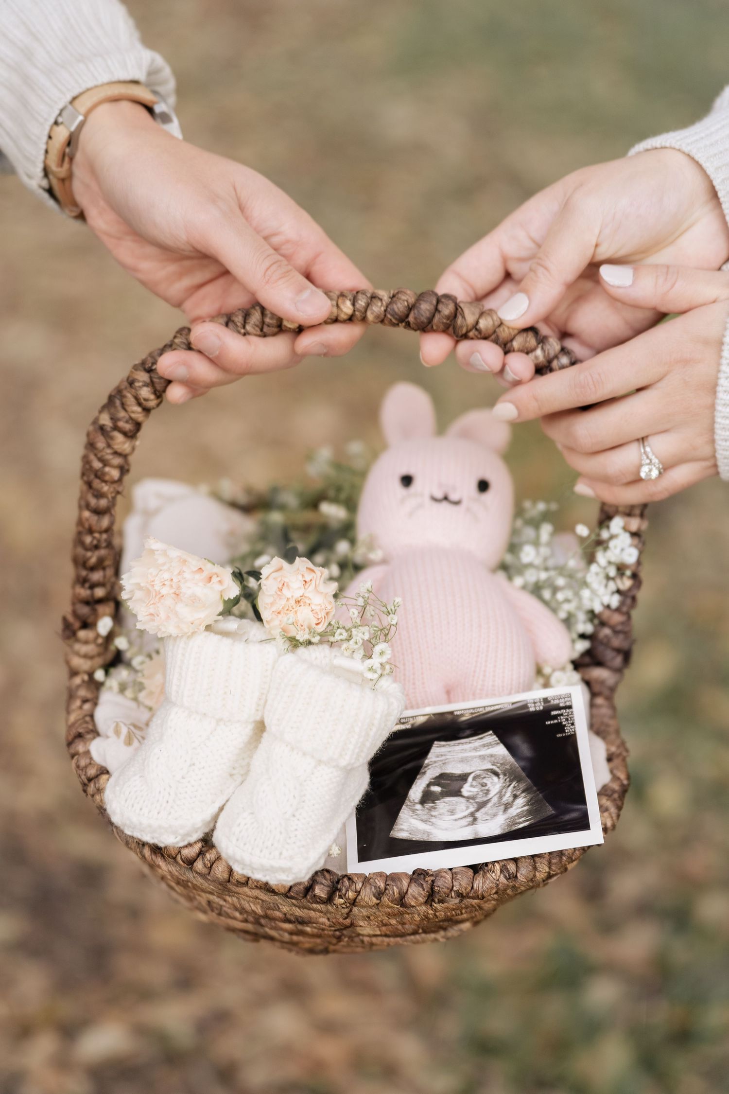 Couple holding wicker basket with pink bunny, ultrasound photo, white booties, and flowers for baby announcement.