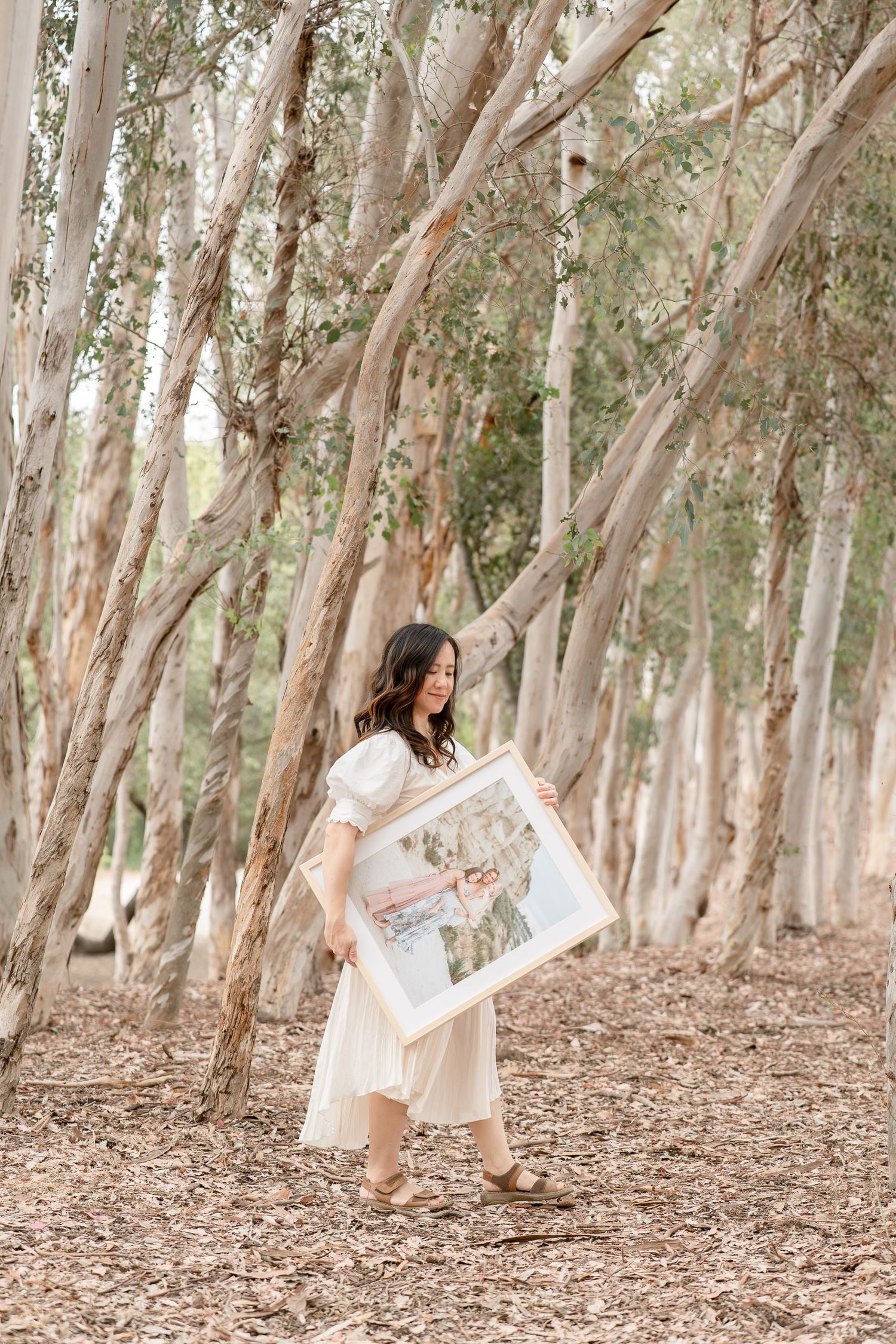 Woman in cream dress holding a framed photo while walking through a eucalyptus forest.