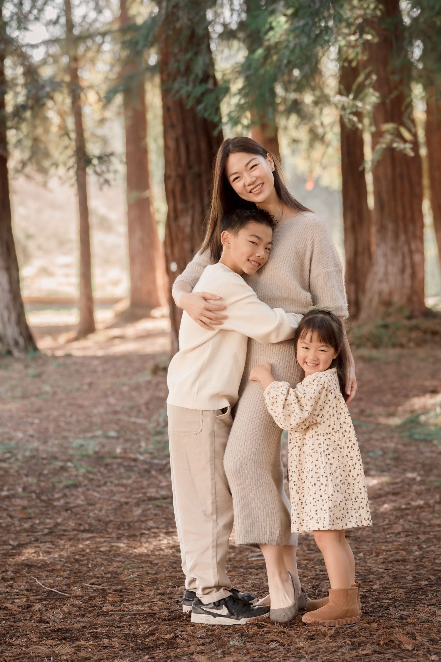 Smiling Asian mother hugging son and daughter in a sunlit forest with tall trees.