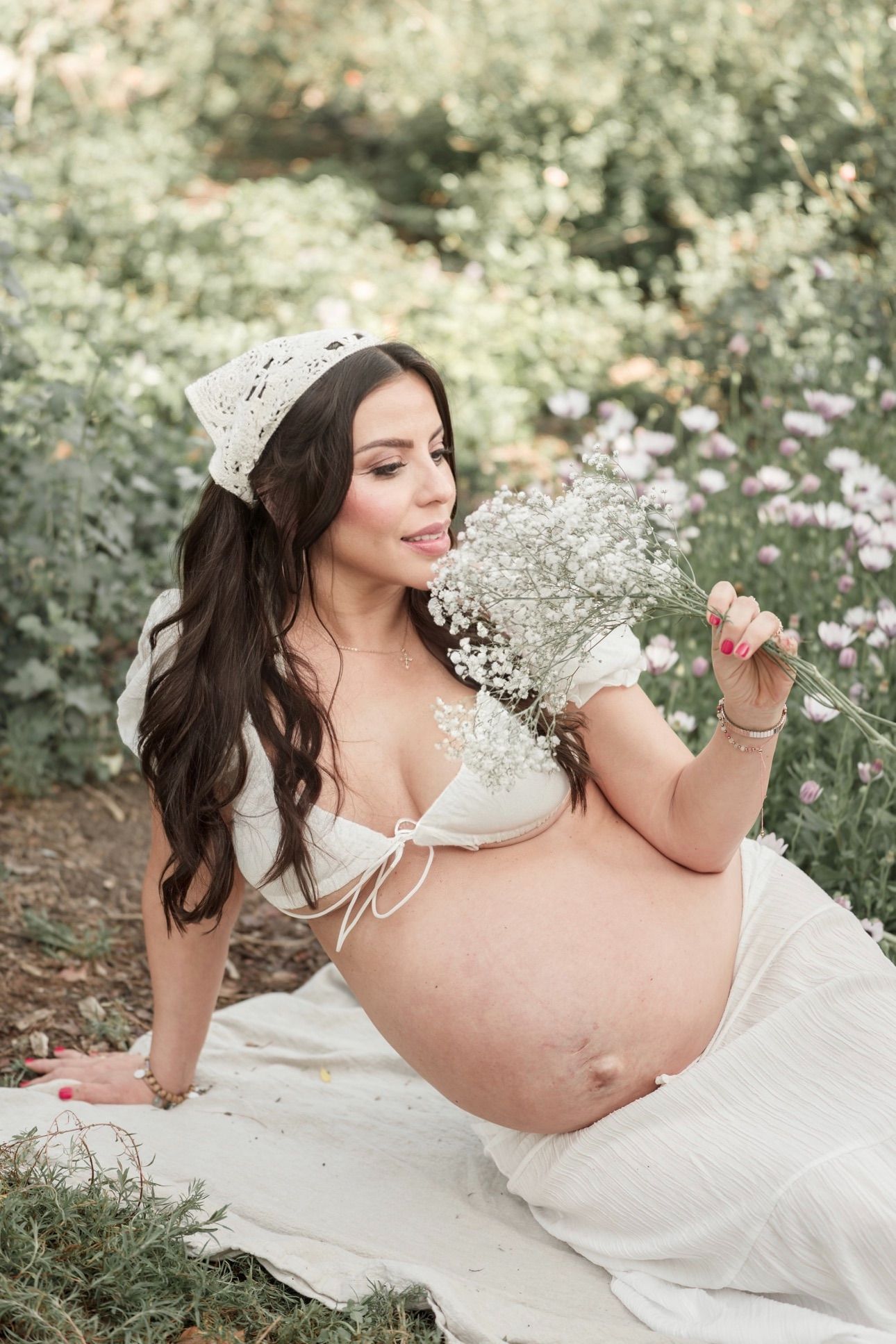Pregnant woman in white bikini top and skirt holds baby's breath flowers outdoors among blooming garden flowers.