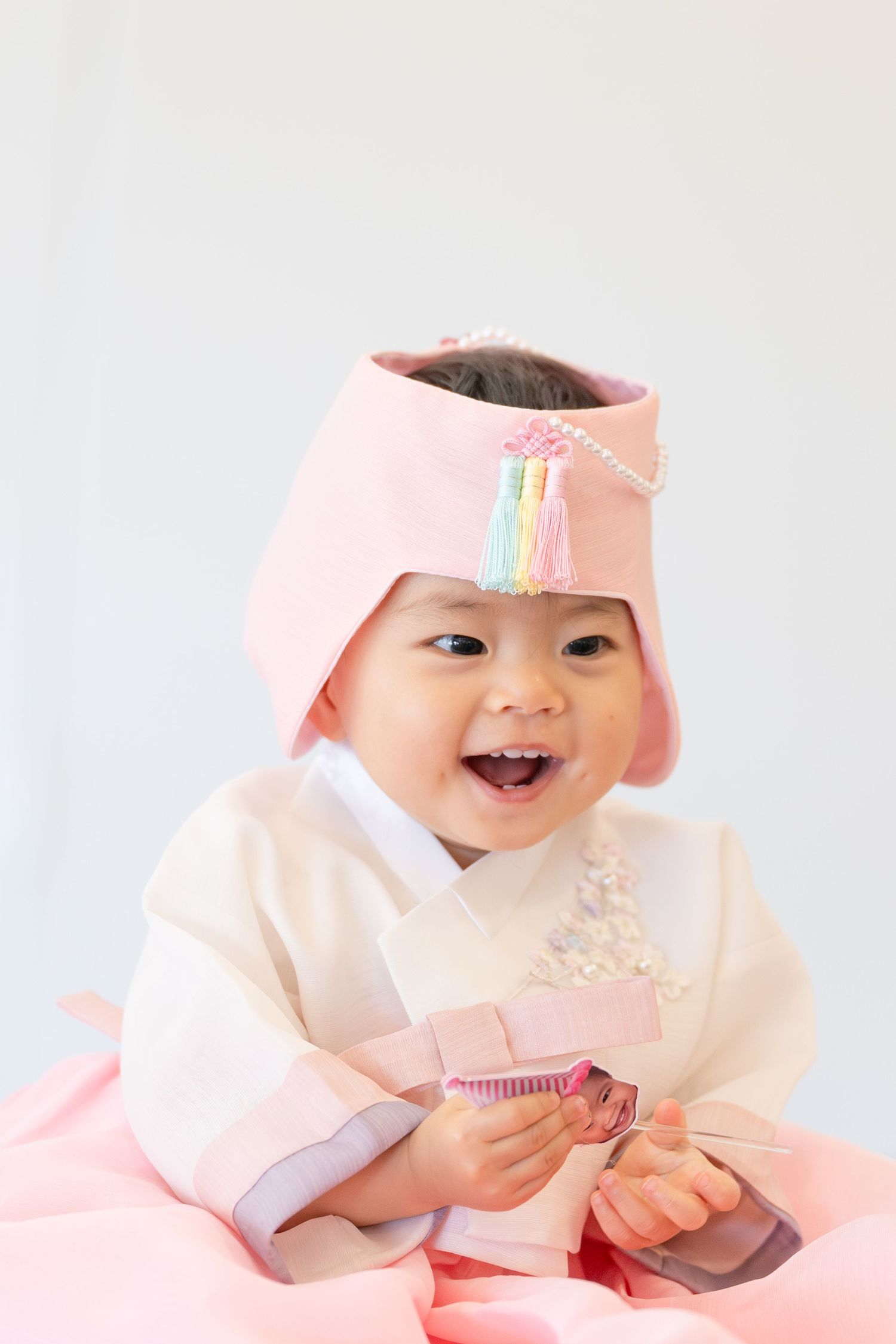 Smiling baby in traditional Korean hanbok with pink hat and colorful tassel for first birthday dol celebration.