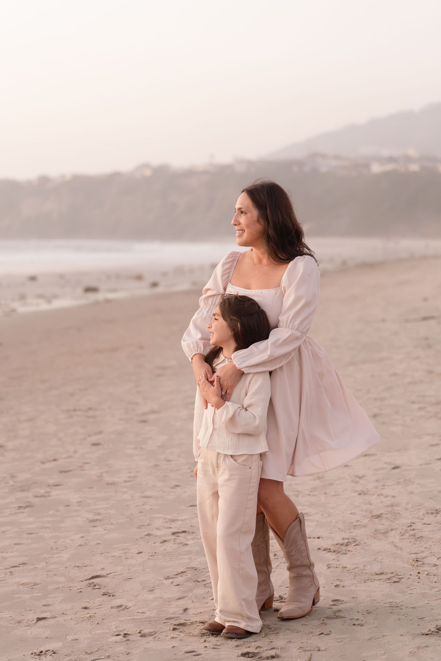 A smiling woman in a white dress and boots hugs a child on a sandy beach at sunset.