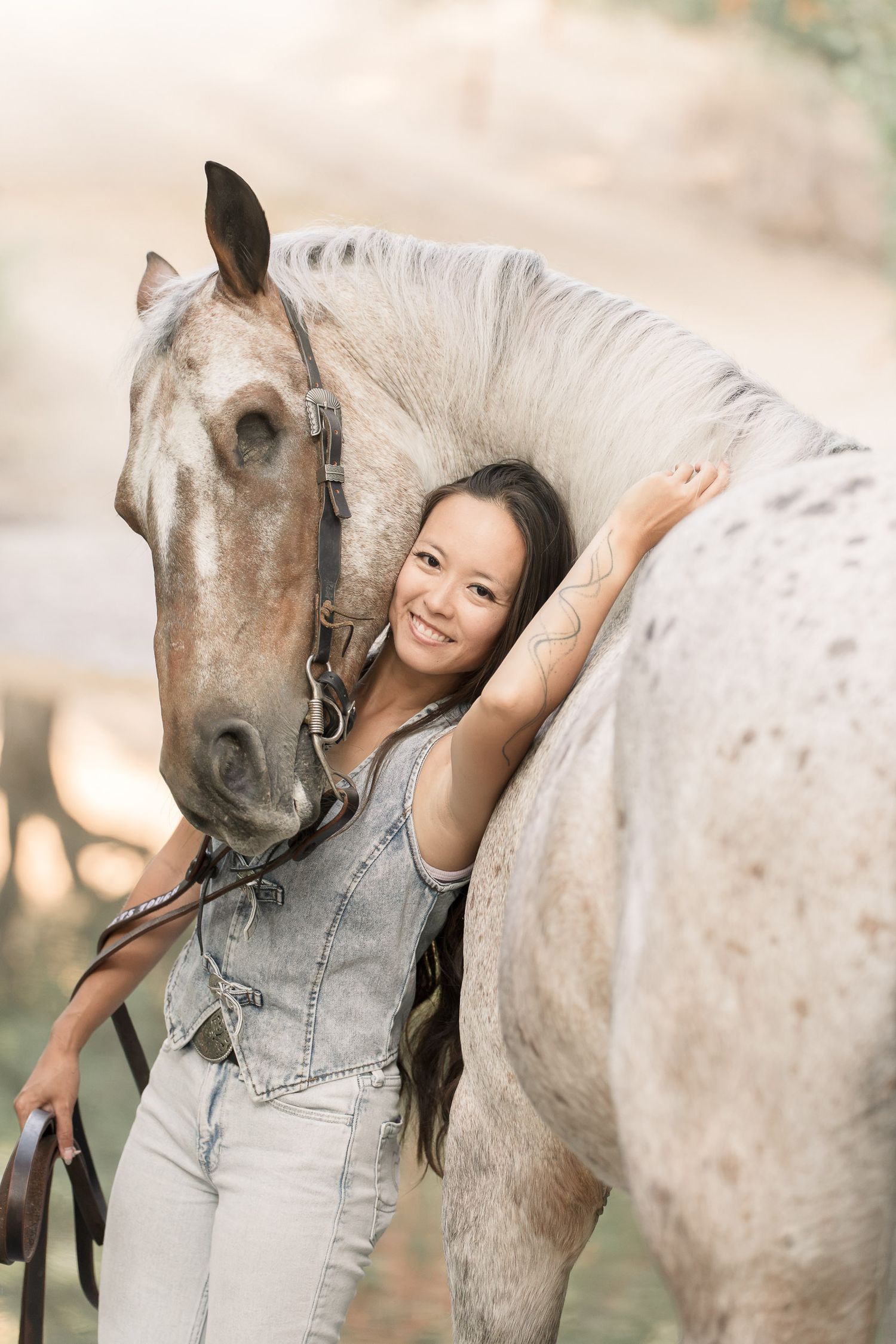 A smiling young woman in denim hugs a dapple gray horse outdoors in warm natural light.