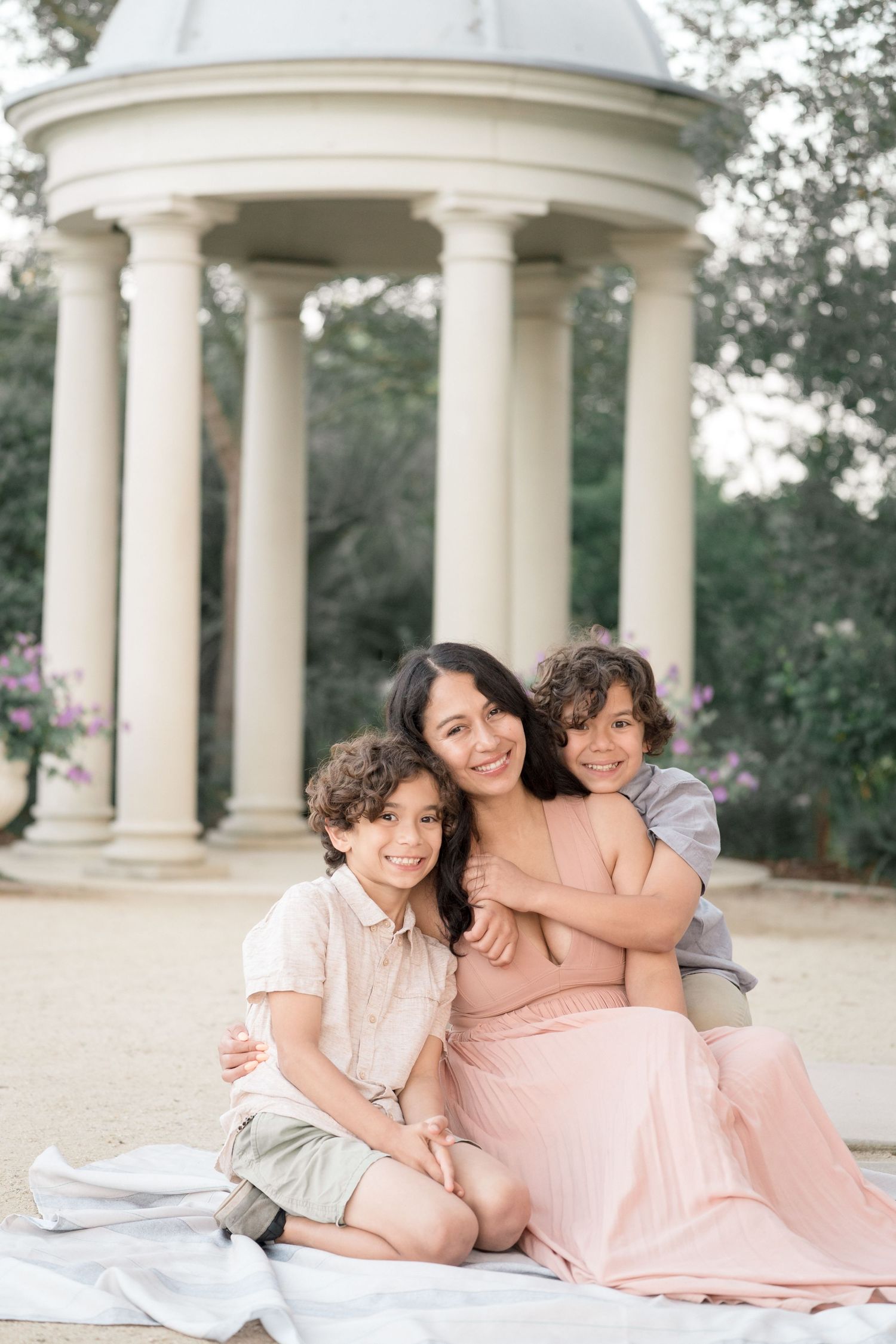 A smiling mother in a pink dress sits on a blanket with her two young sons before a white rotunda.