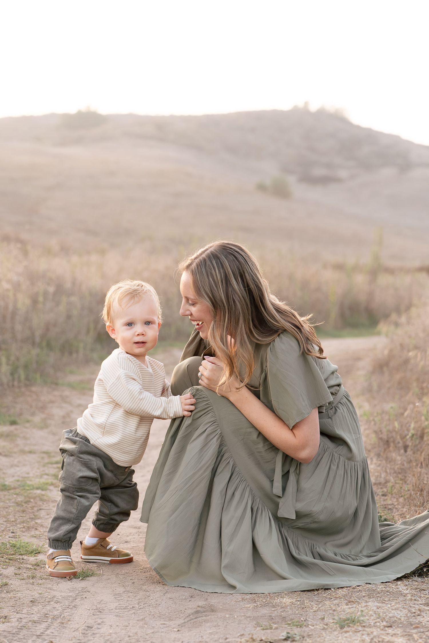 A mother in a sage green dress crouches on a dirt path, smiling at her toddler son in a sunlit field.