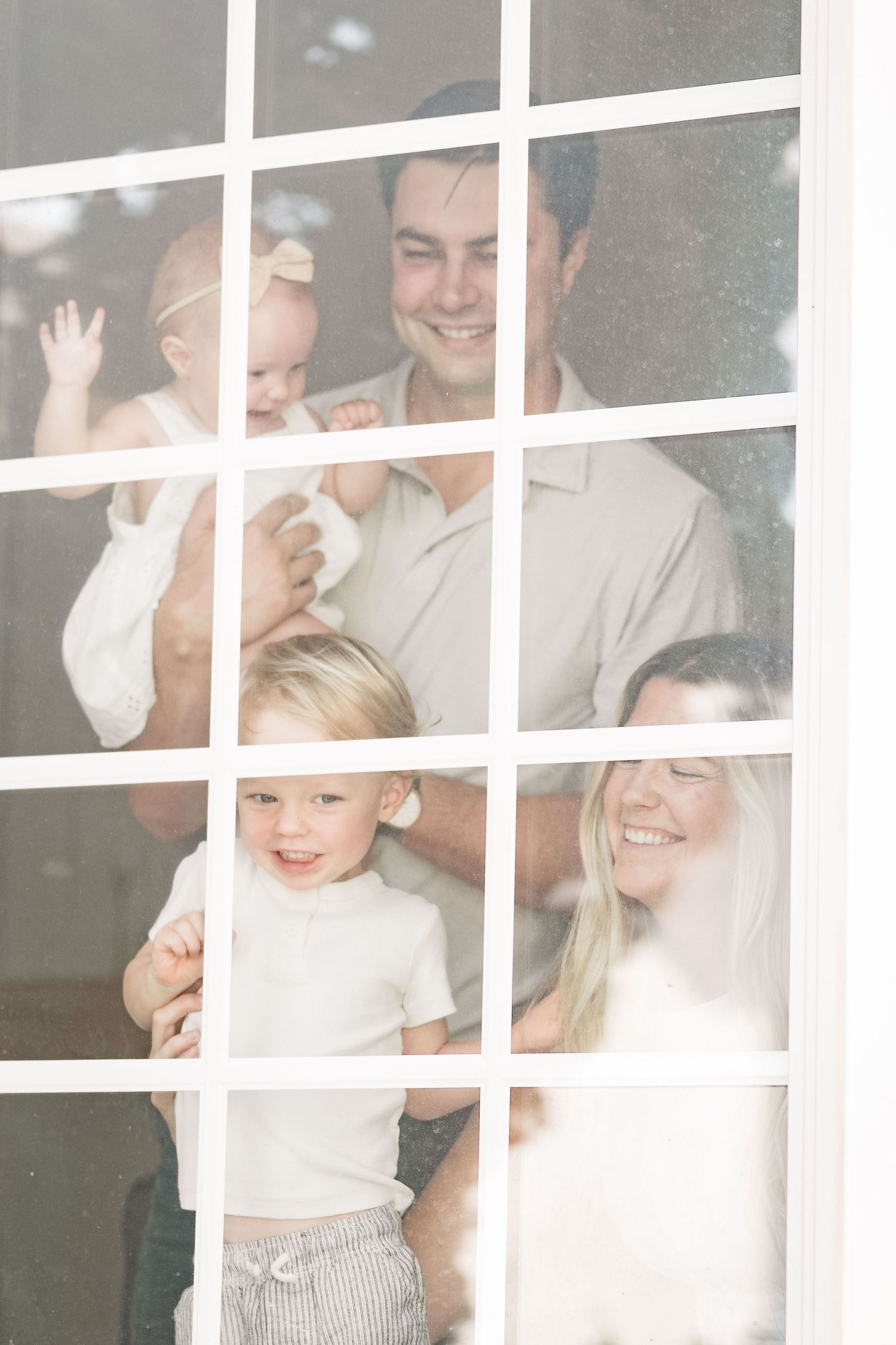 Happy family of four smiling through a white-paned window, parents holding two young children.