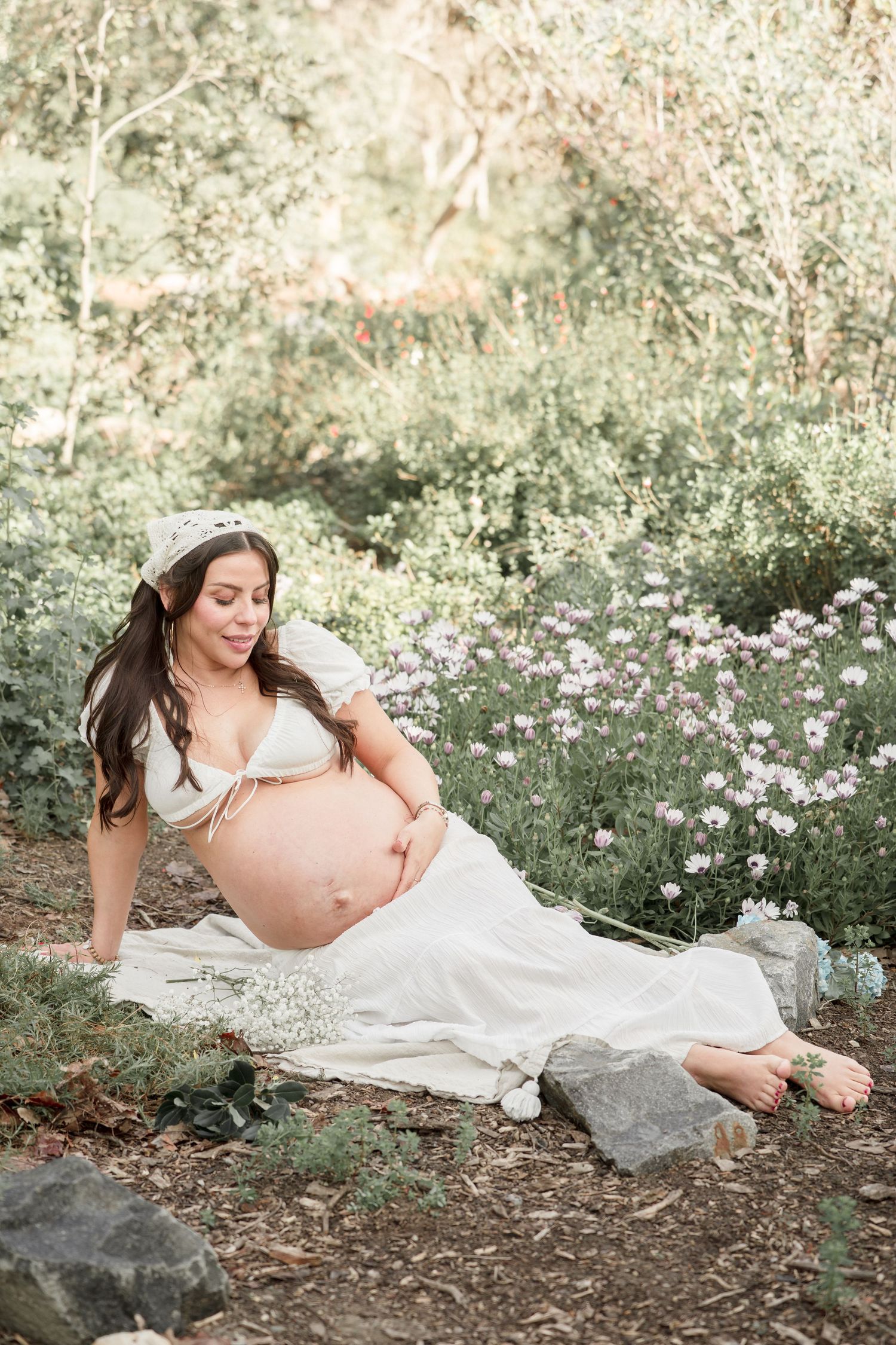 Pregnant woman in white dress sitting outdoors among wildflowers, cradling her bare baby bump, wearing a floral crown.
