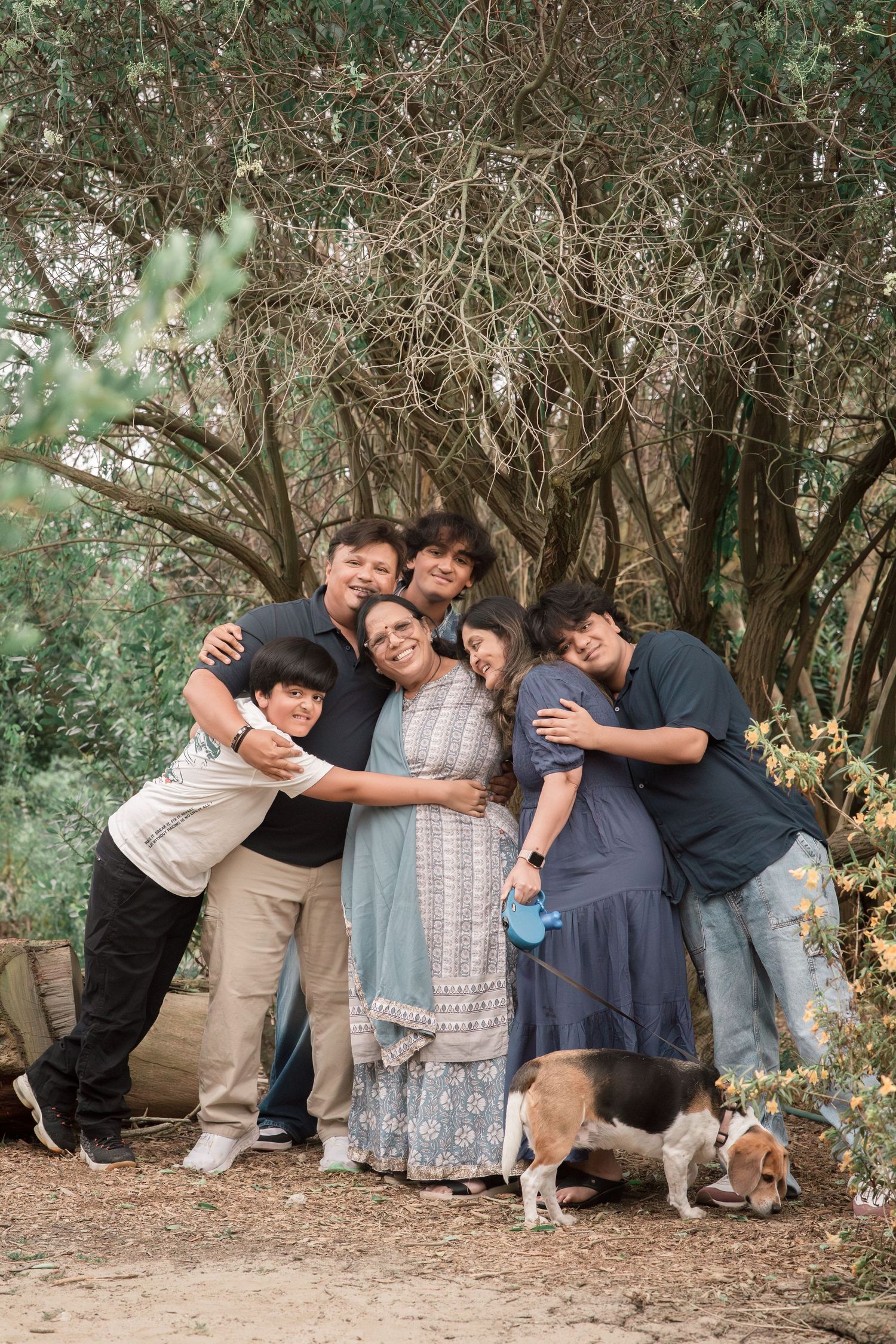 Large Indian family of six hugging warmly outdoors with a beagle dog nearby.