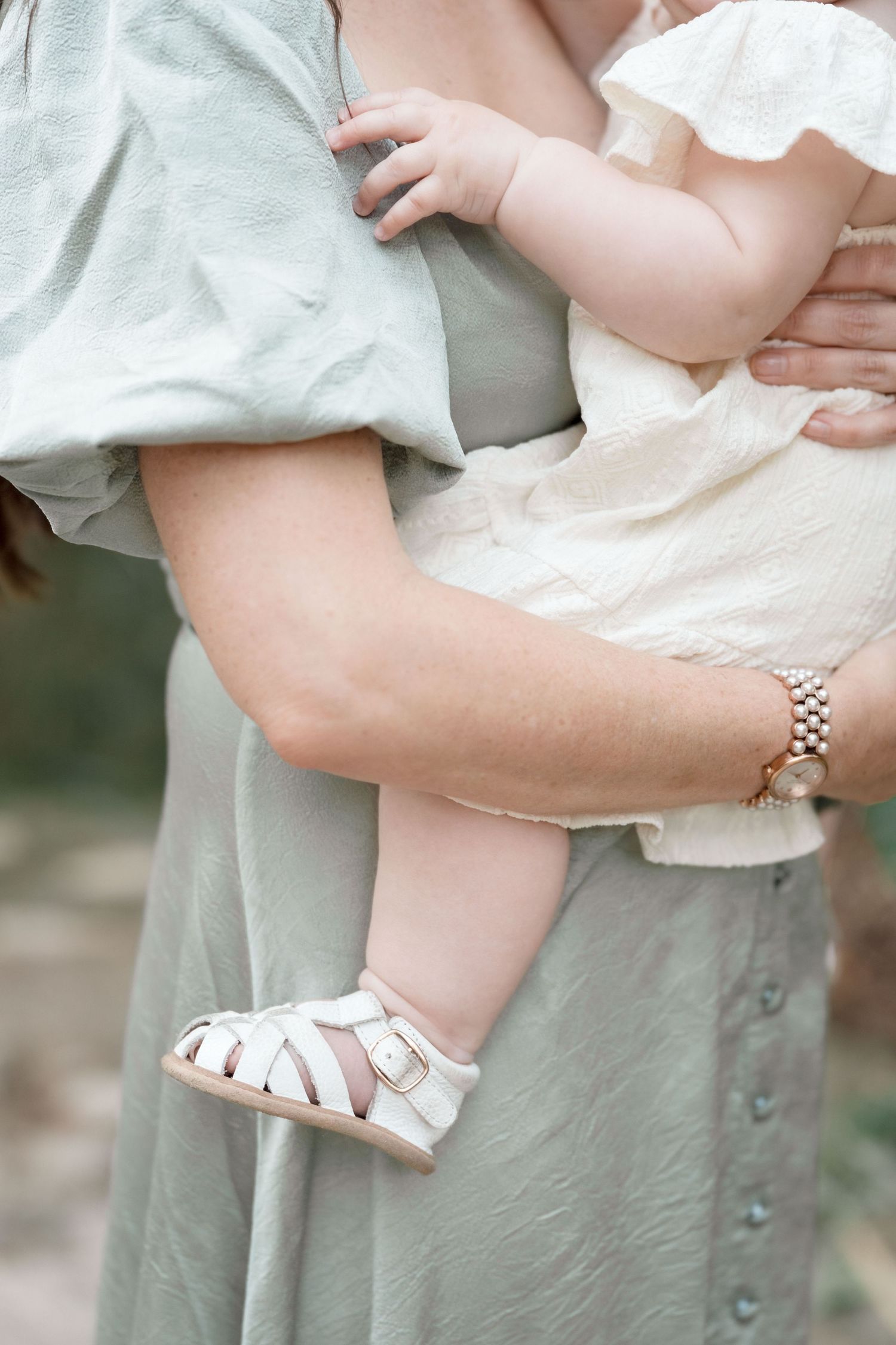 Close-up of baby in cream dress with white sandals being held by mother in sage green dress.