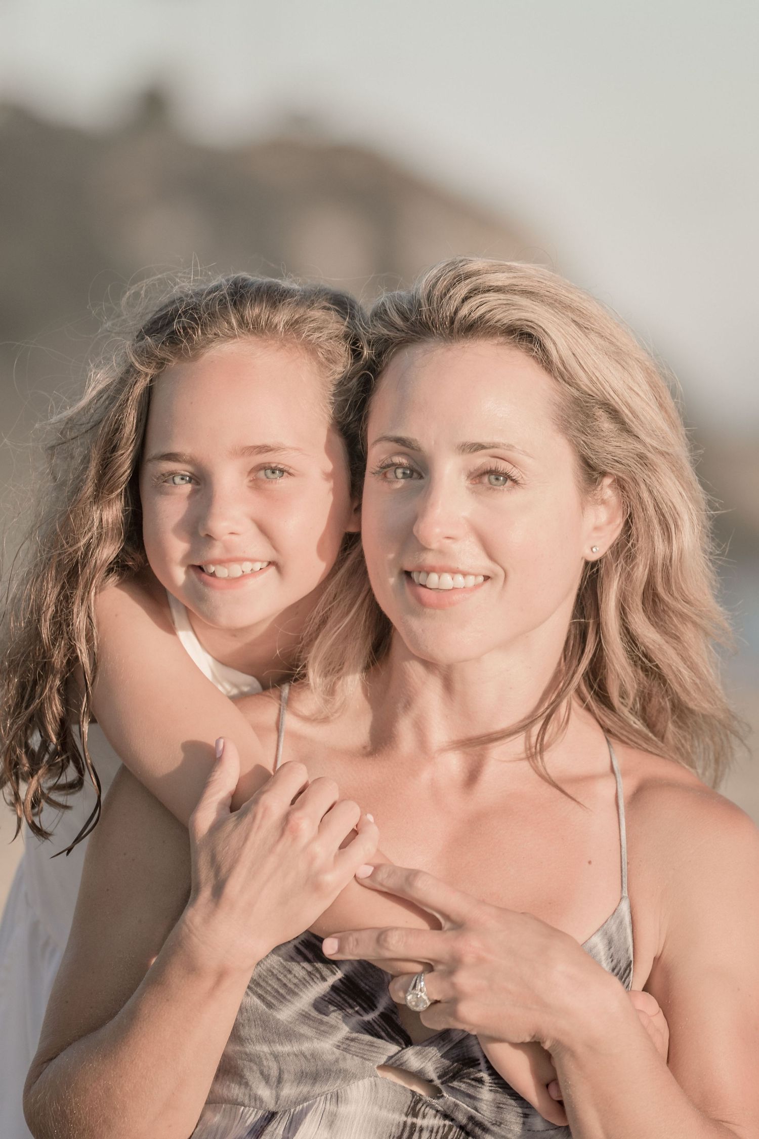 Smiling blonde mother and daughter share a tender embrace on a sun-drenched beach at golden hour.