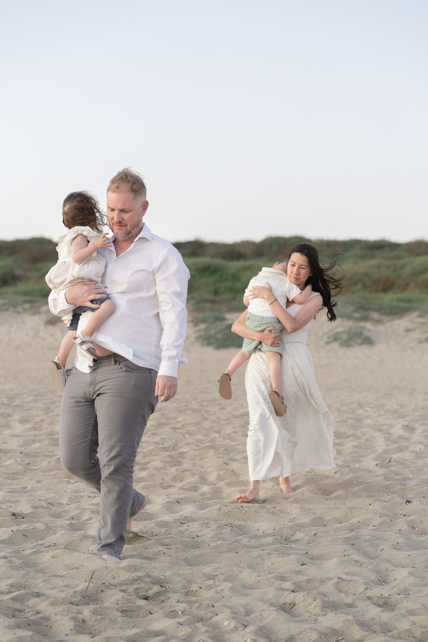 A family of four walks on a sandy beach, parents each carrying a young child in soft, light-colored clothing.