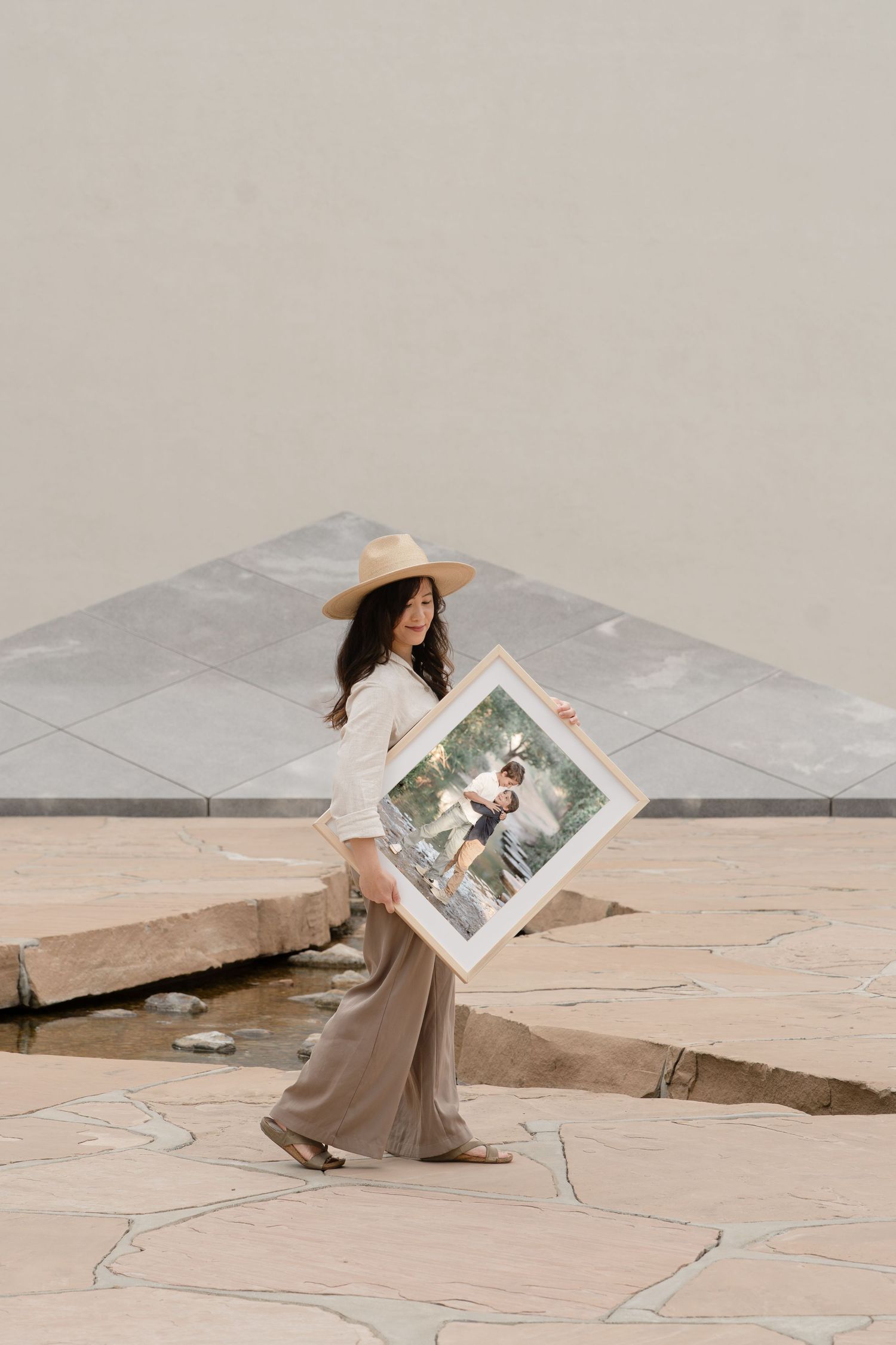 Woman in straw hat and wide-leg pants holding a large framed photo outdoors on stone pavement.