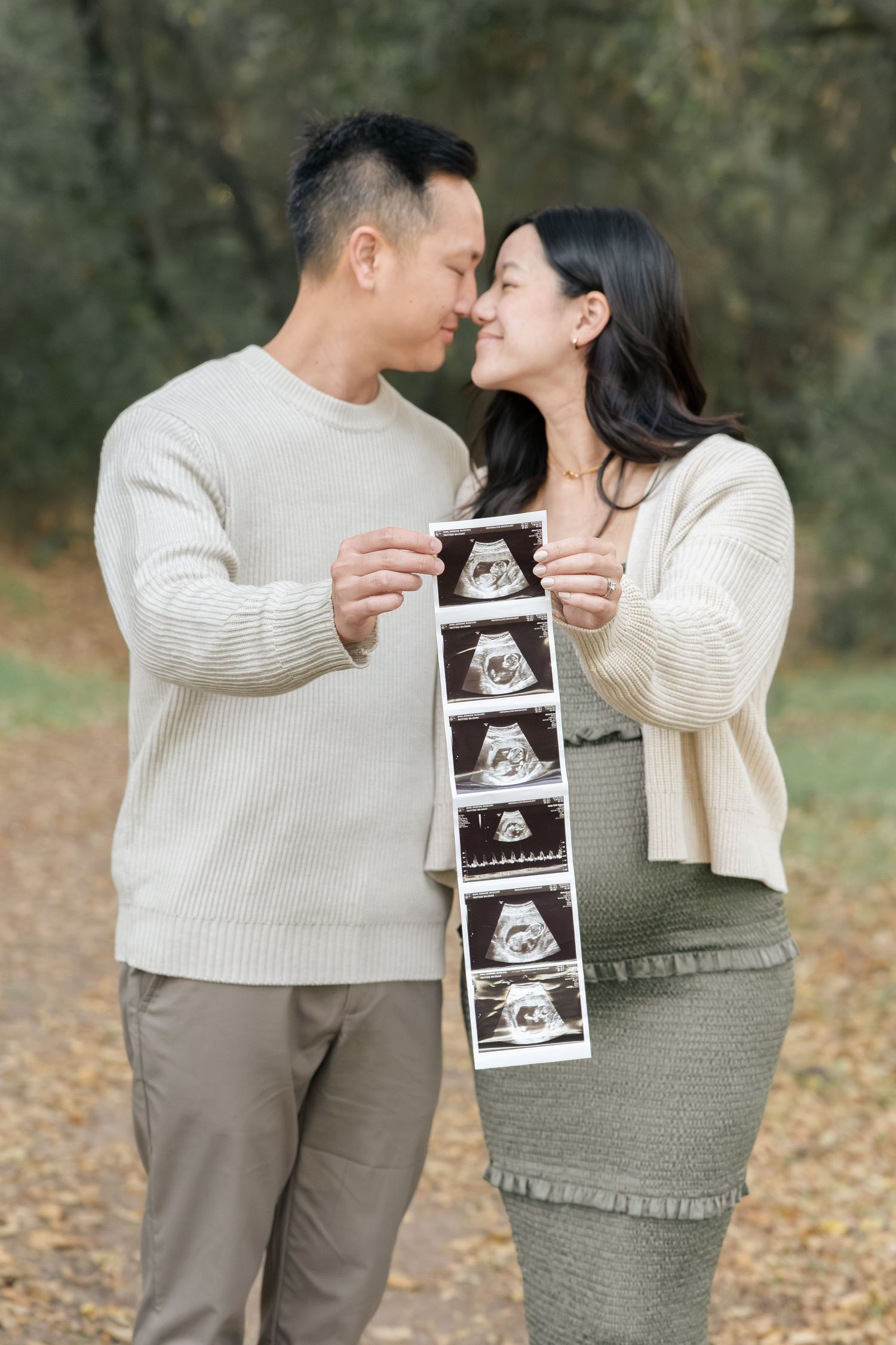 Asian couple nearly kissing while holding ultrasound strip photos in autumn park setting.