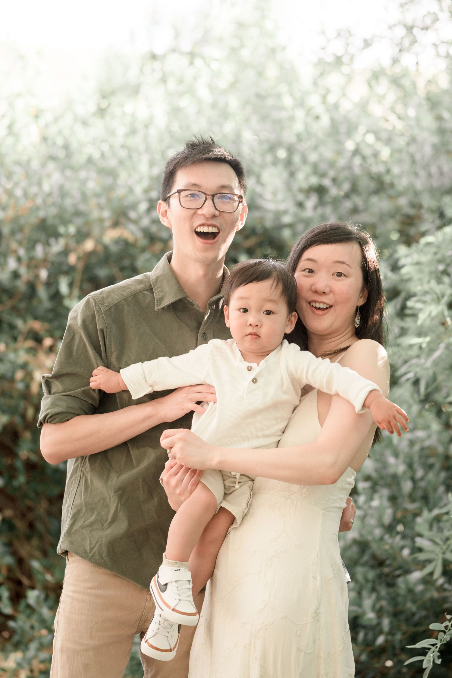 Happy Asian family portrait with parents and toddler smiling outdoors among lush green foliage.