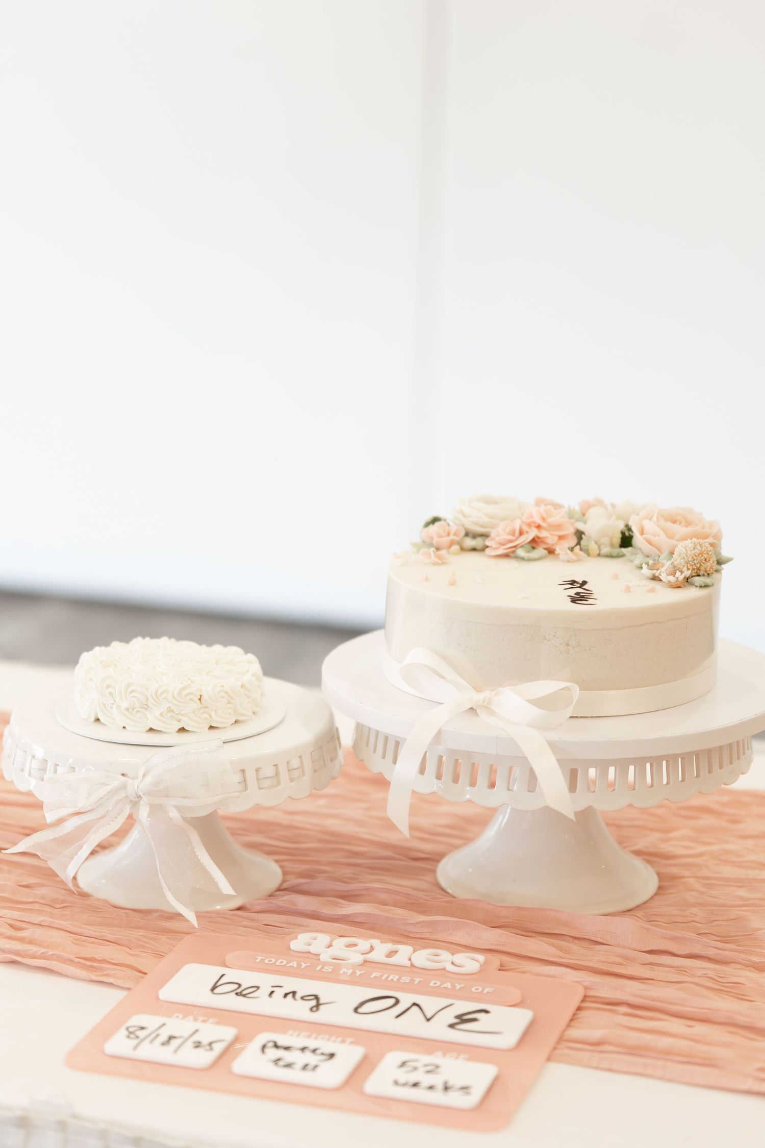 Two elegant white cakes on decorative stands displayed on a peach table with a being ONE milestone card.