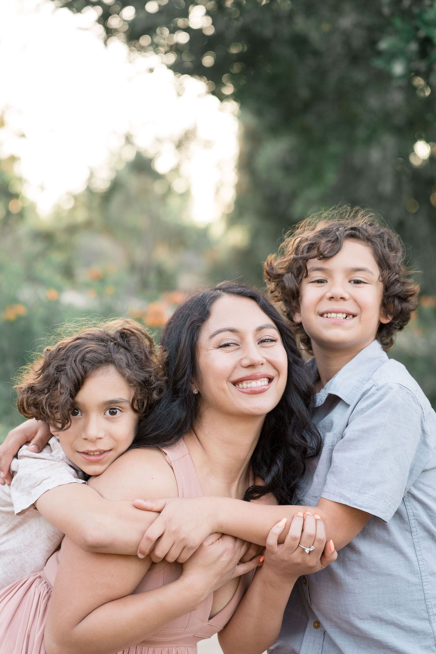 Joyful dark-haired mother smiling while two curly-haired sons embrace her outdoors in natural light.