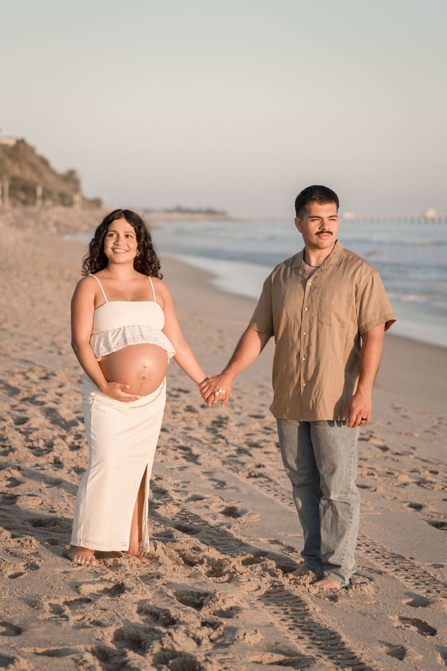 Pregnant woman in white dress holding hands with mustachioed partner walking on sandy beach at sunset.