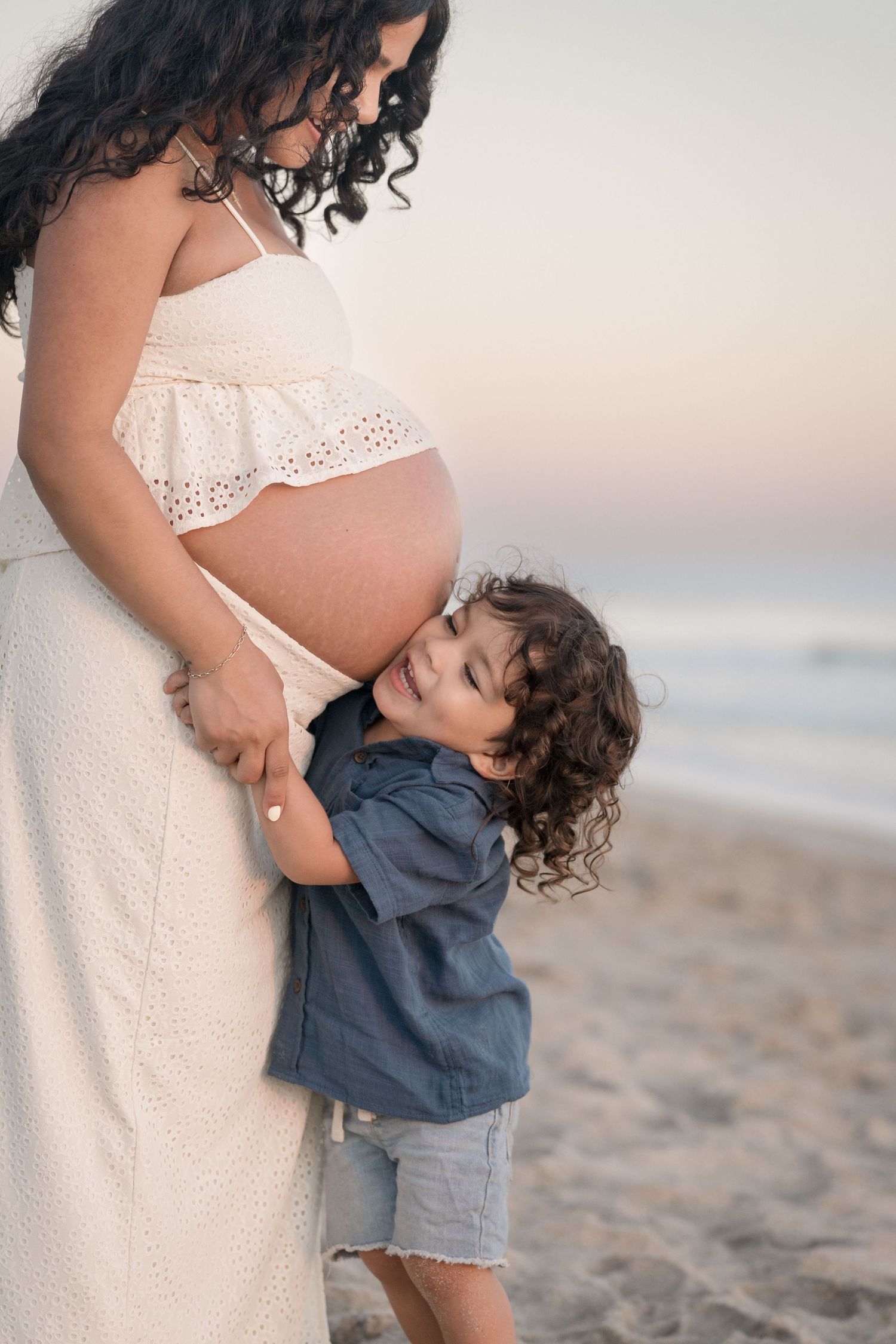 Happy child embracing pregnant mothers belly on sandy beach at dusk.