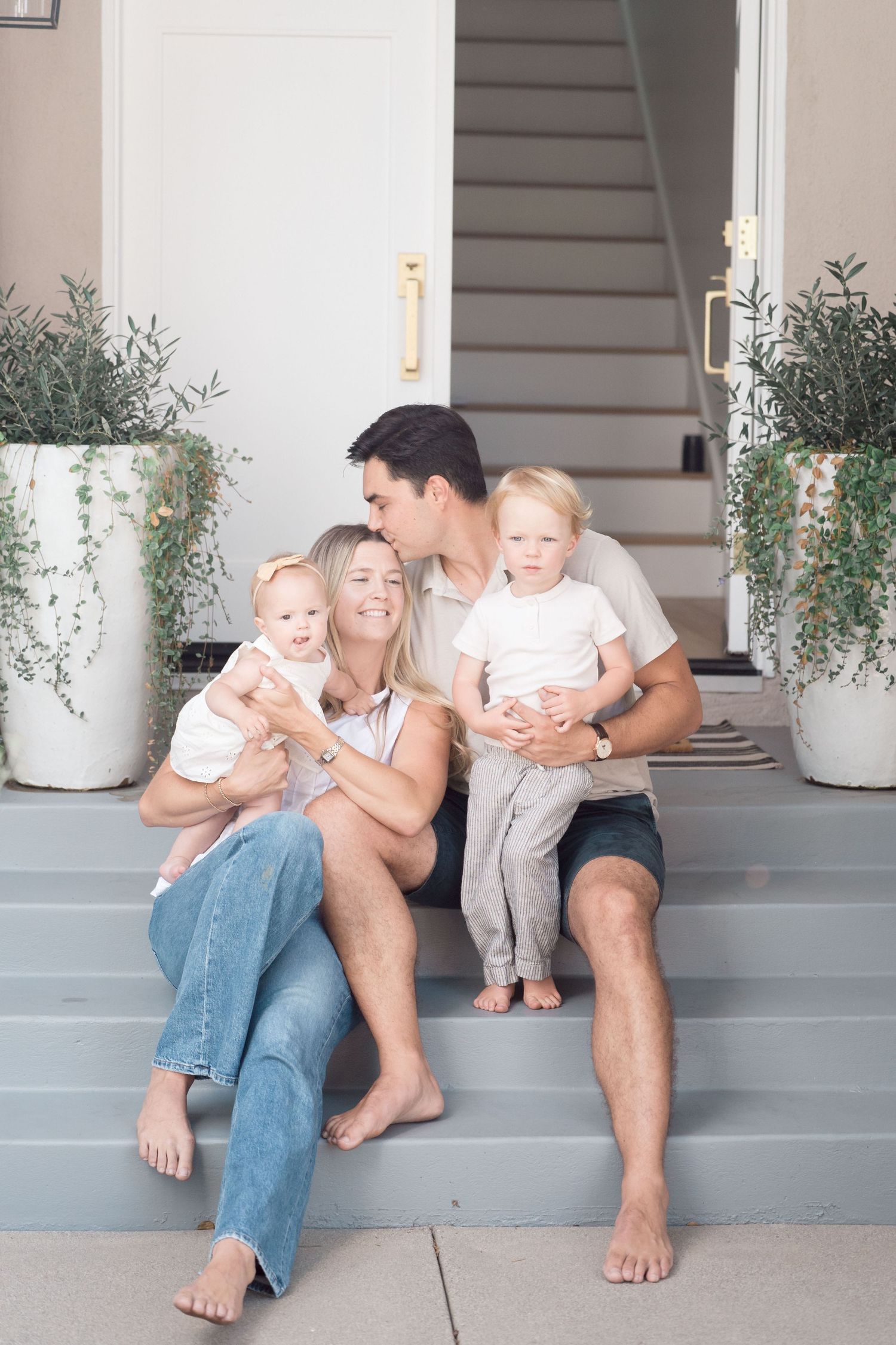 Family of four sitting on front porch steps with parents kissing beside two toddlers.