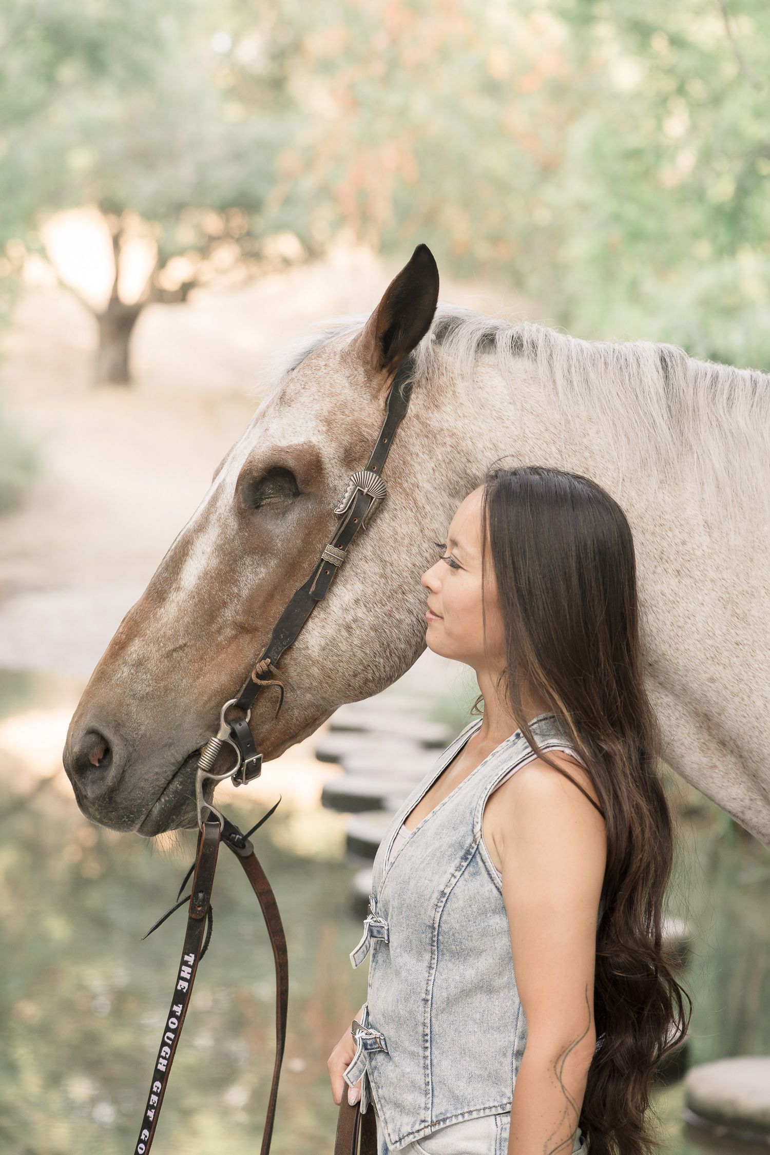 Young woman in denim overalls sharing a tender moment with a gray horse outdoors among trees.