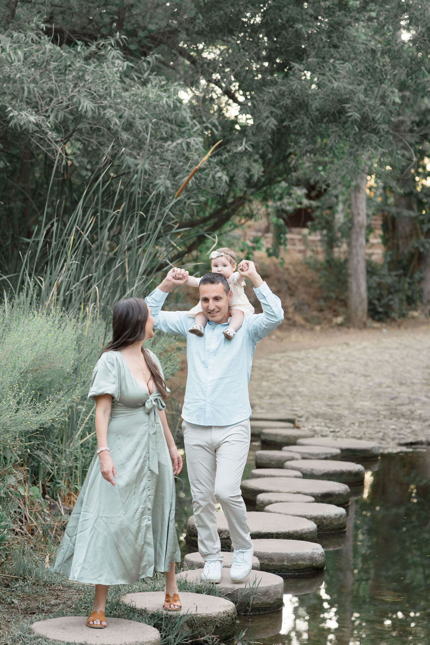 Joyful family walks on stepping stones as father carries baby on shoulders beside a pond.