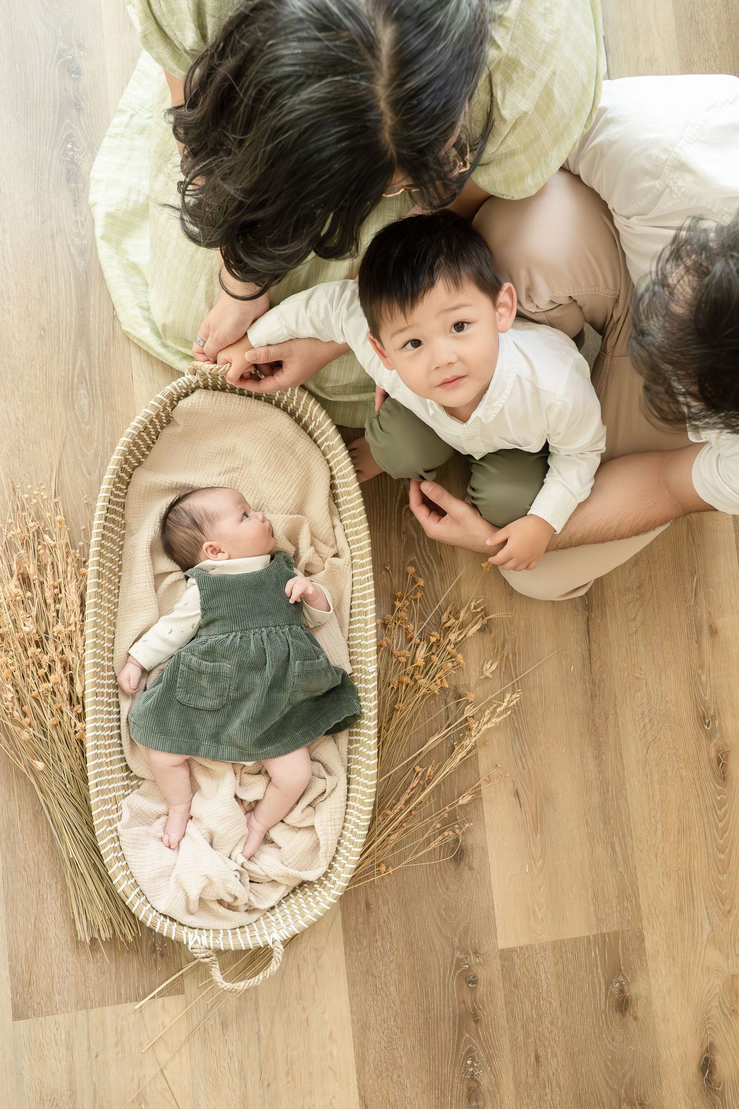 An aerial view of a family with a toddler and newborn baby in a wicker Moses basket on a wooden floor.