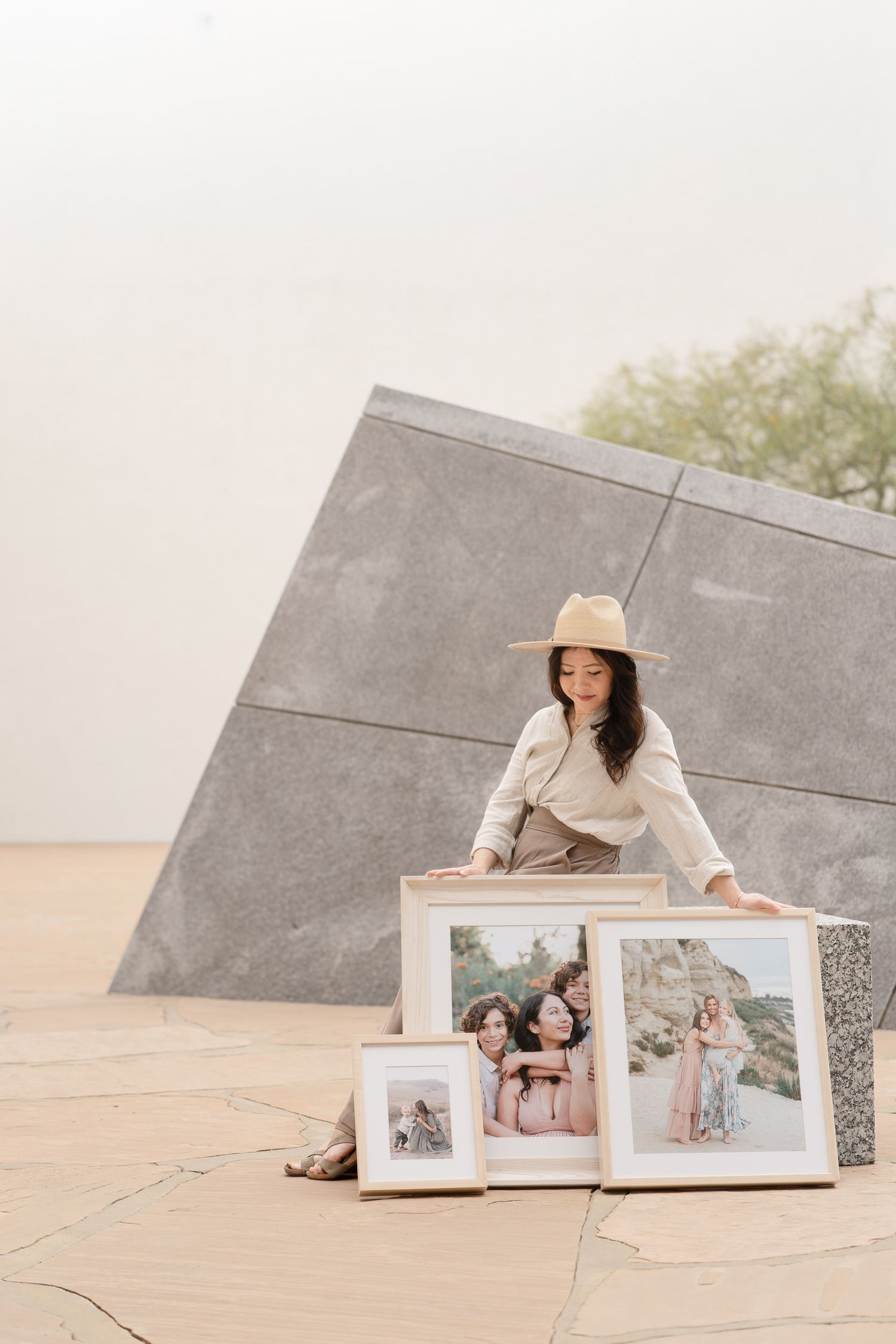 Woman in hat displays framed family portrait photos against a modern stone sculpture outdoors.