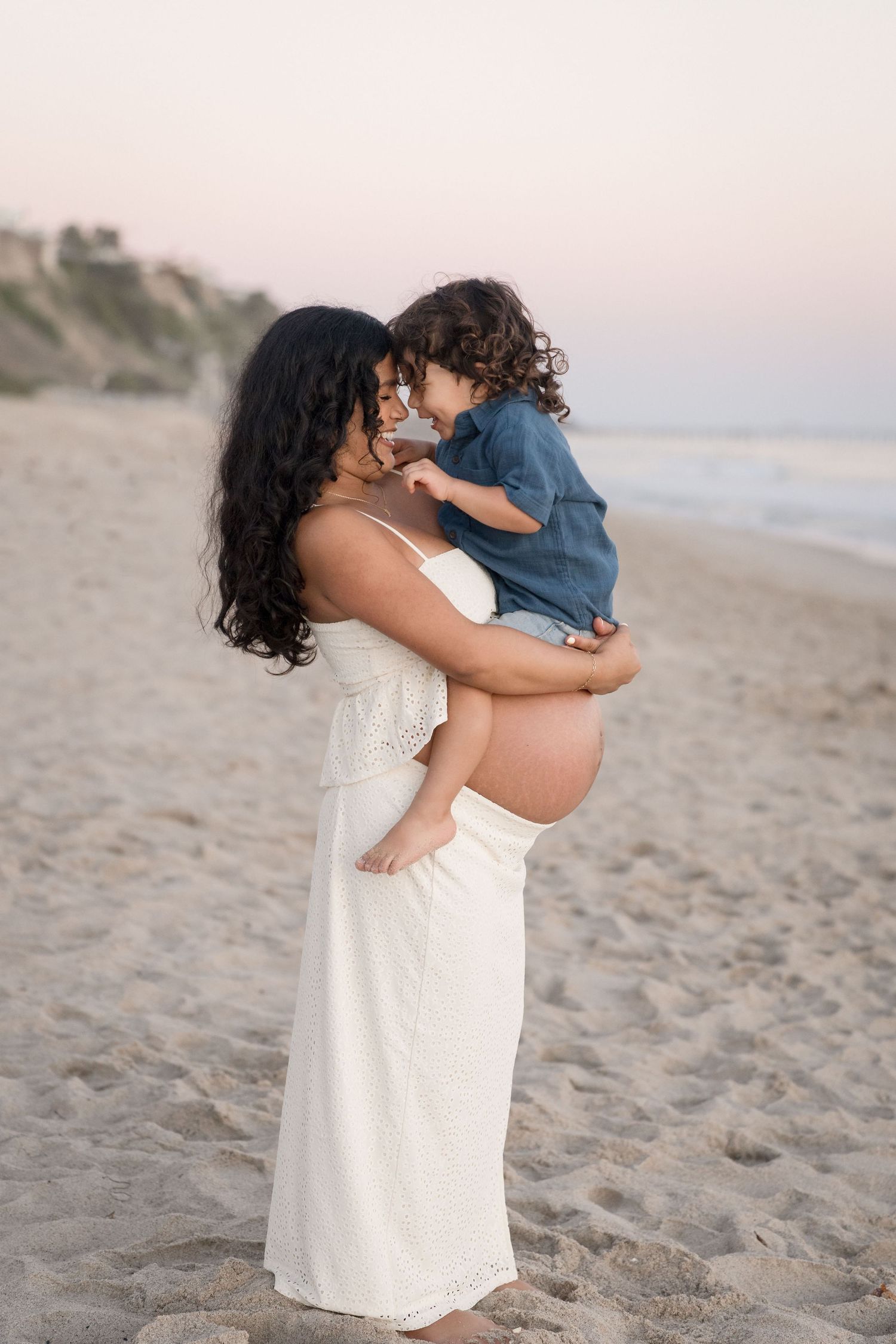 Pregnant woman in white maxi dress holds toddler child face-to-face on a serene beach at golden hour.