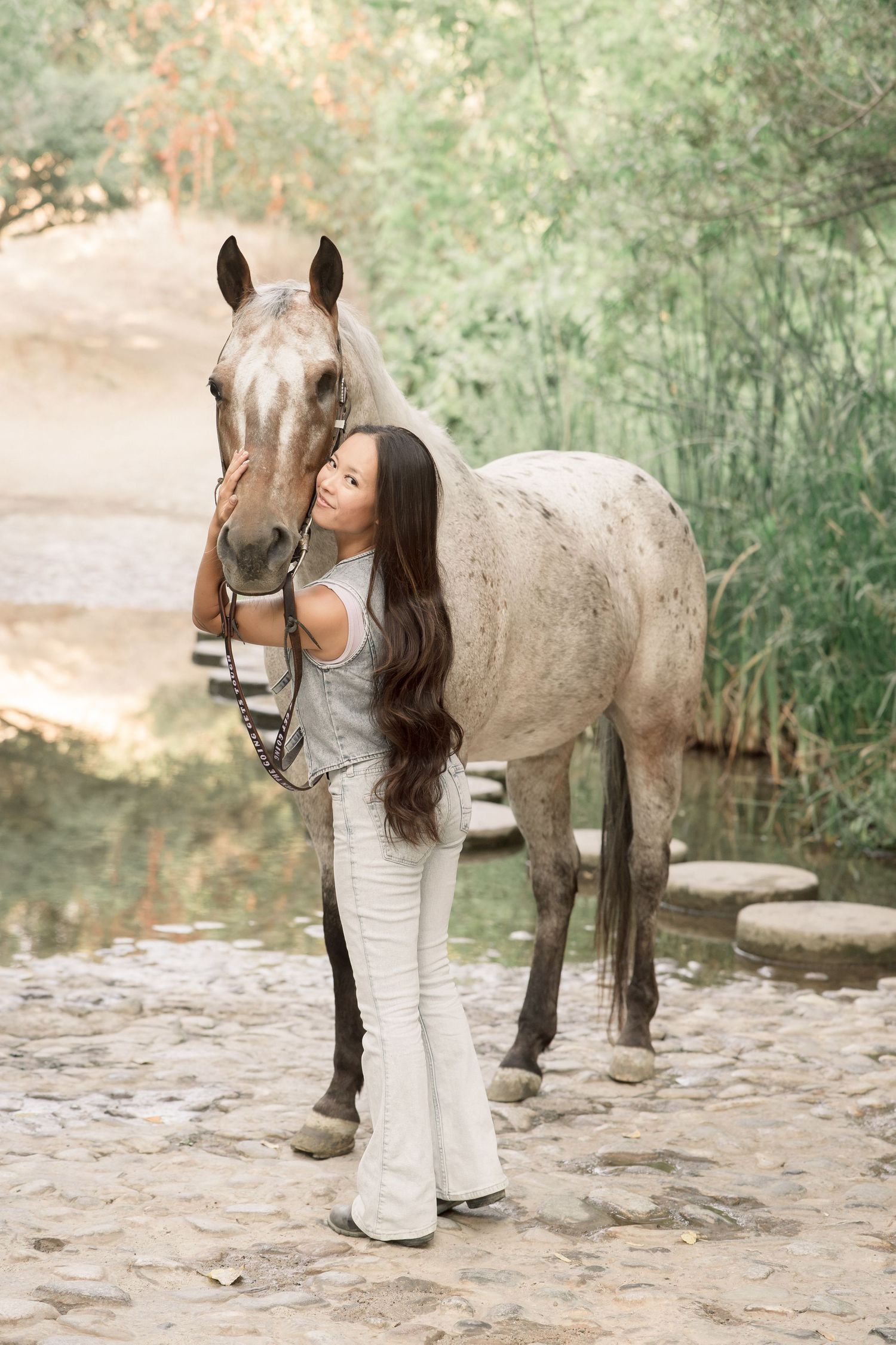 A smiling woman with long dark hair in white flared jeans affectionately embraces a gray speckled horse outdoors.
