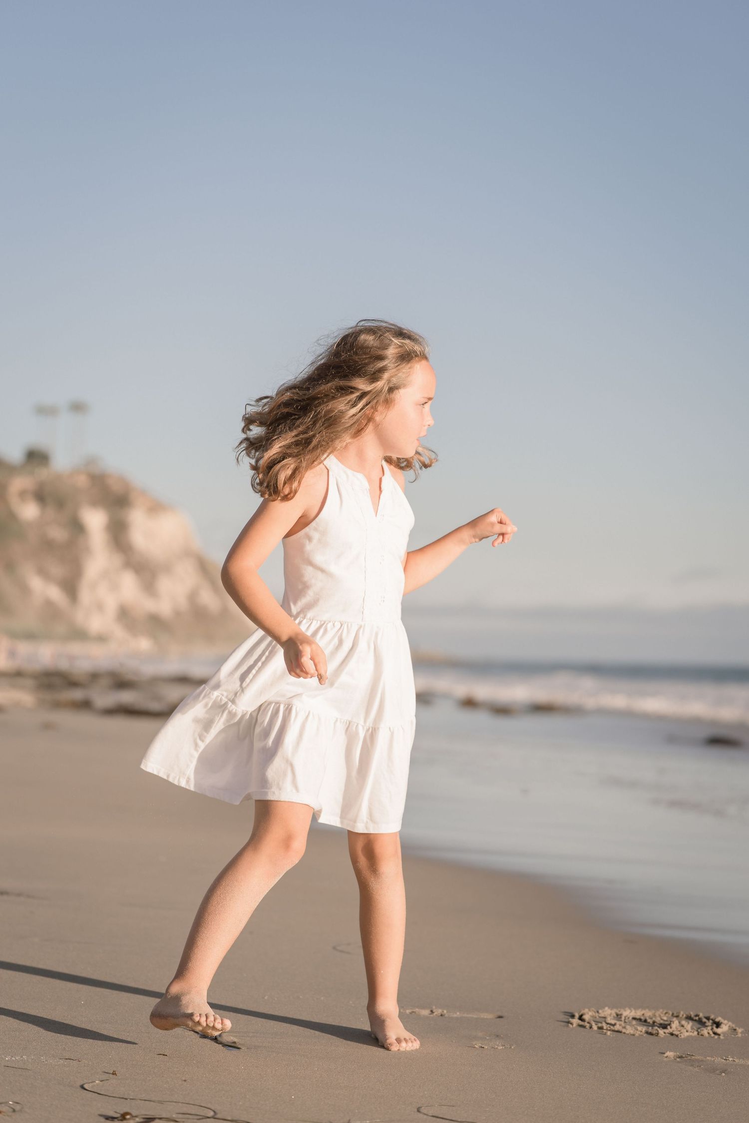 A young girl in a white dress runs barefoot along a sunny beach with ocean waves in the background.