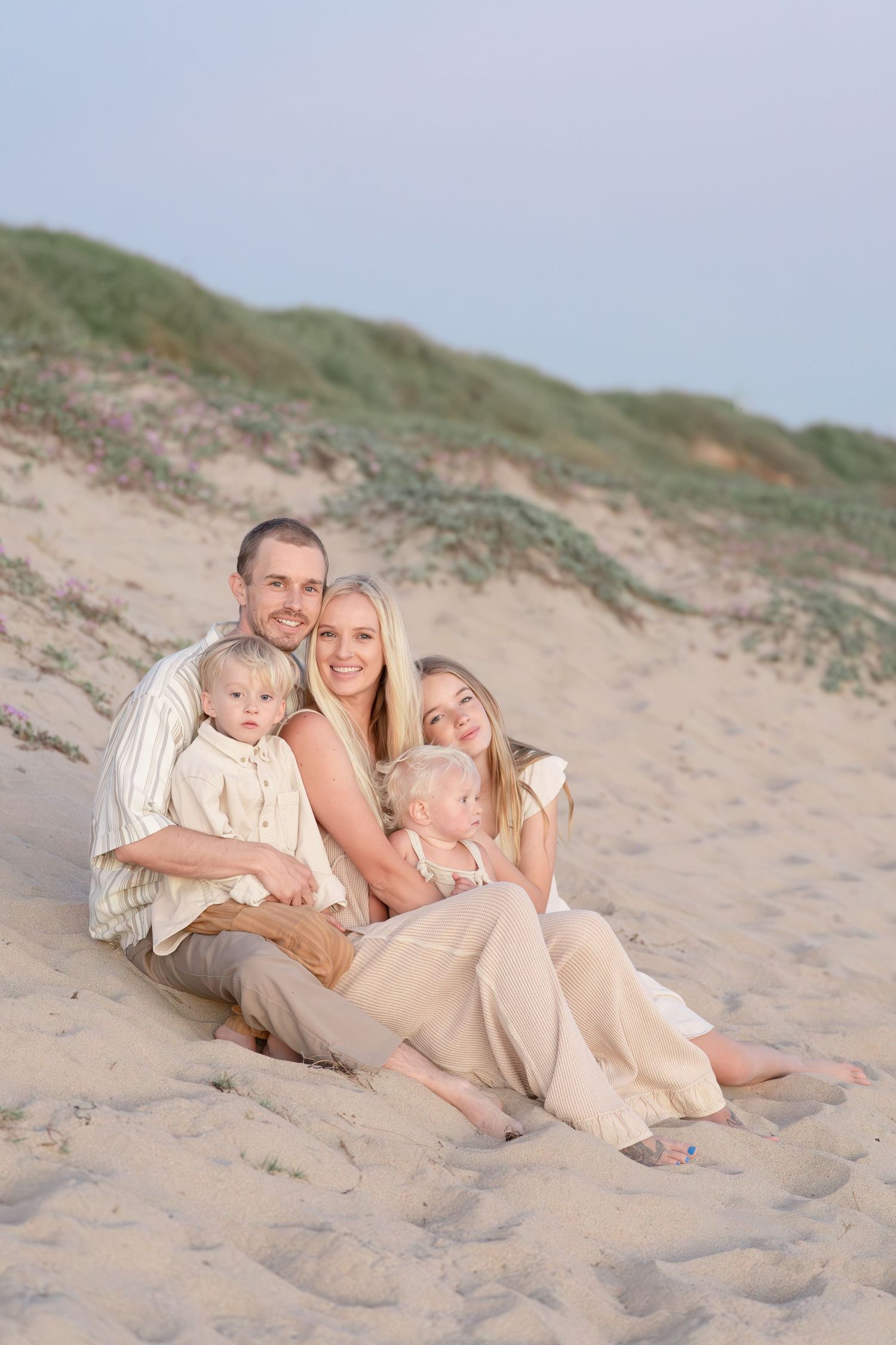 A family of five sits together on sandy beach dunes, smiling warmly in coordinating beige outfits.