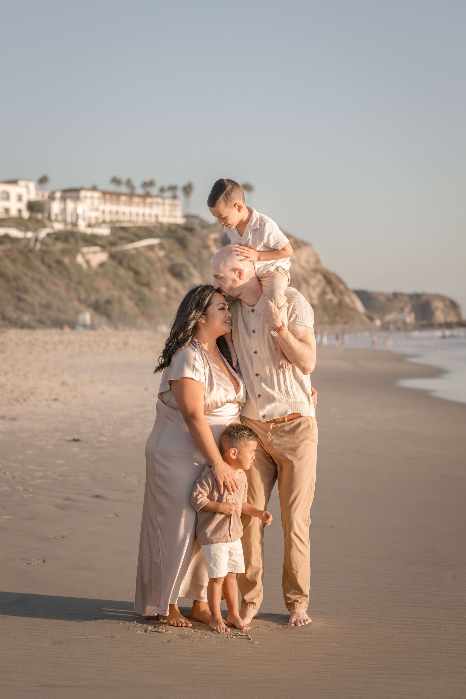 Family of four posing together on a beach at sunset with a cliffside resort in the background.