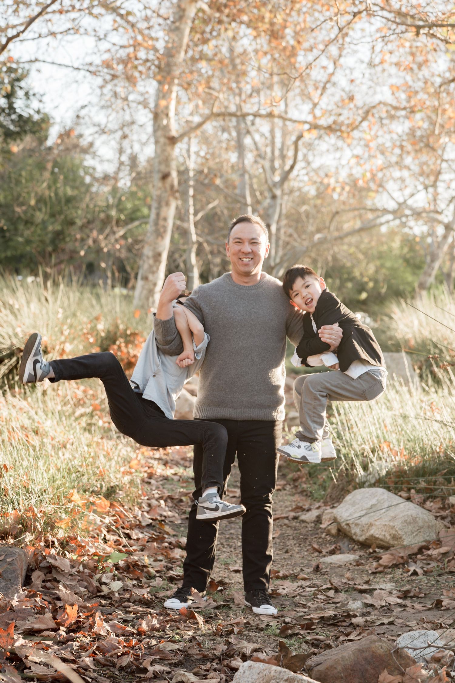 A smiling man playfully holds two laughing children in an autumn park setting with golden foliage.