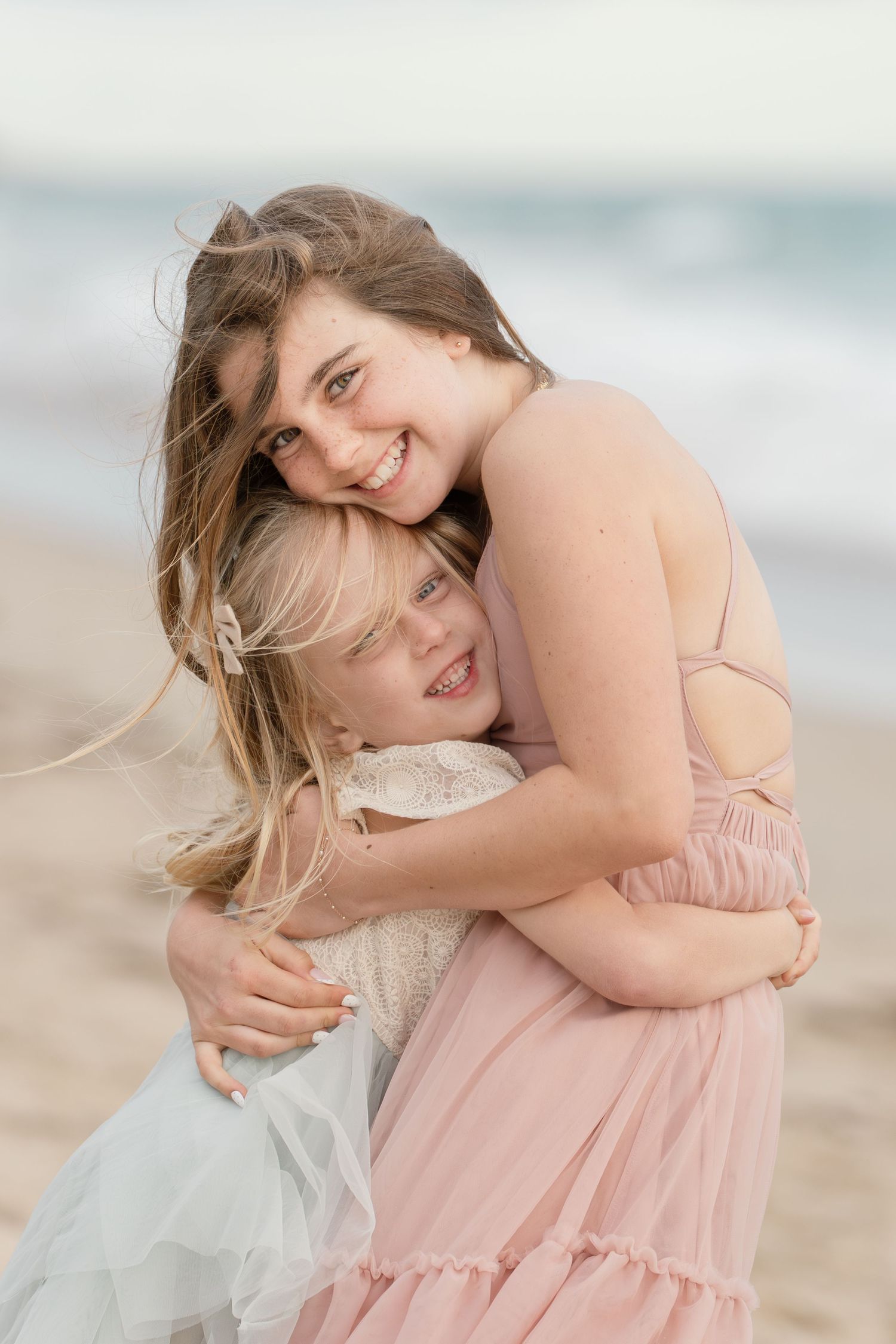 A smiling mother in a pink dress hugs her young blonde daughter on a sandy beach.