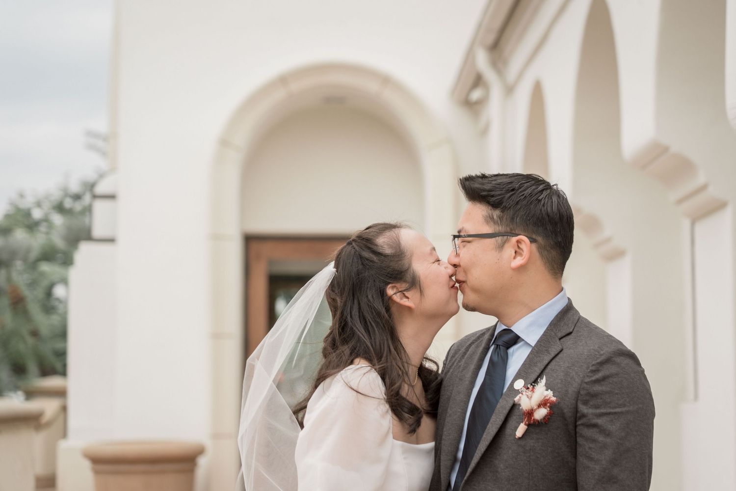 Newlywed couple sharing a romantic kiss outdoors near white arched building on wedding day.