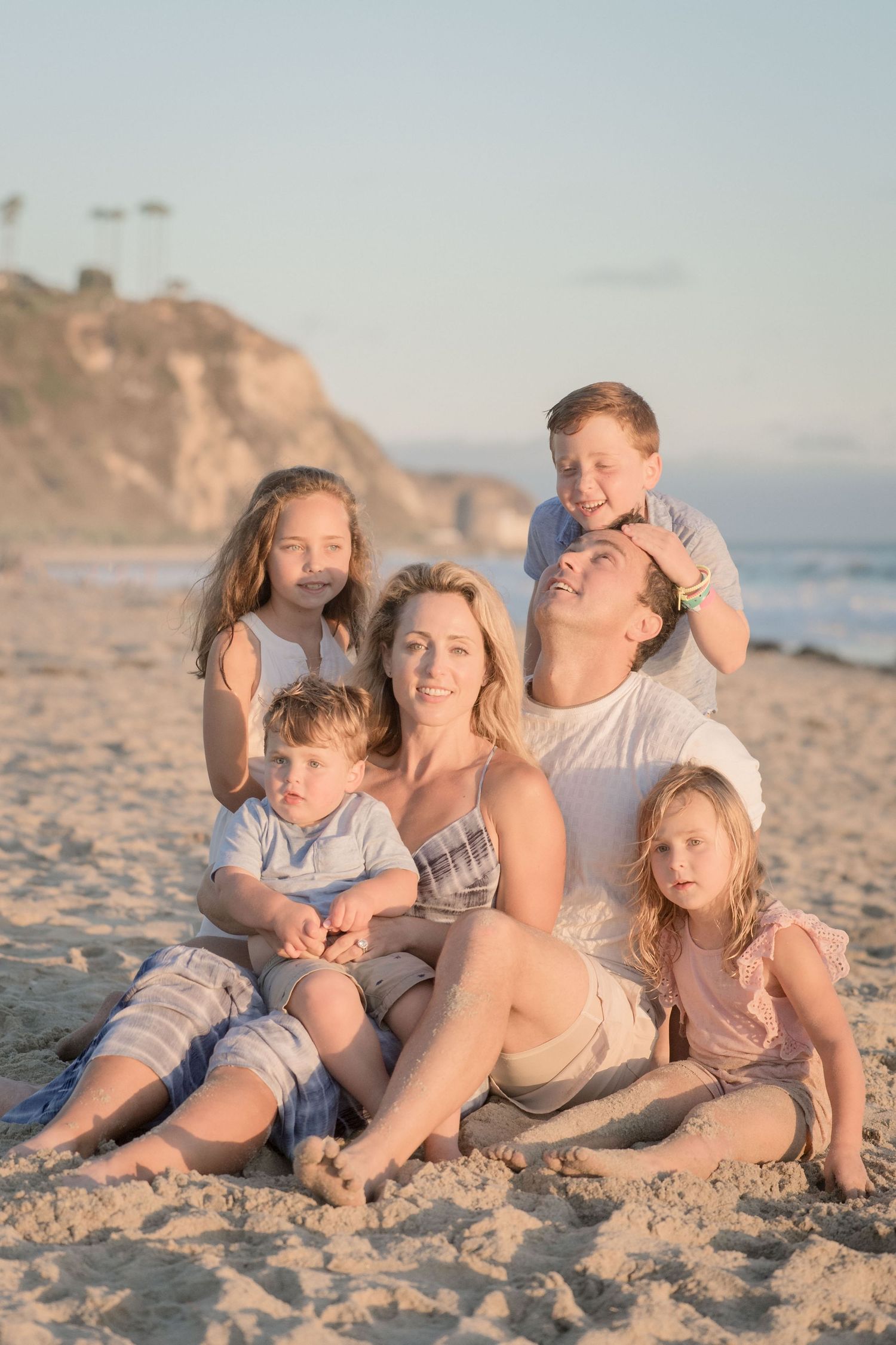 Family of six sitting on beach at sunset with rocky coastal cliffs in background.
