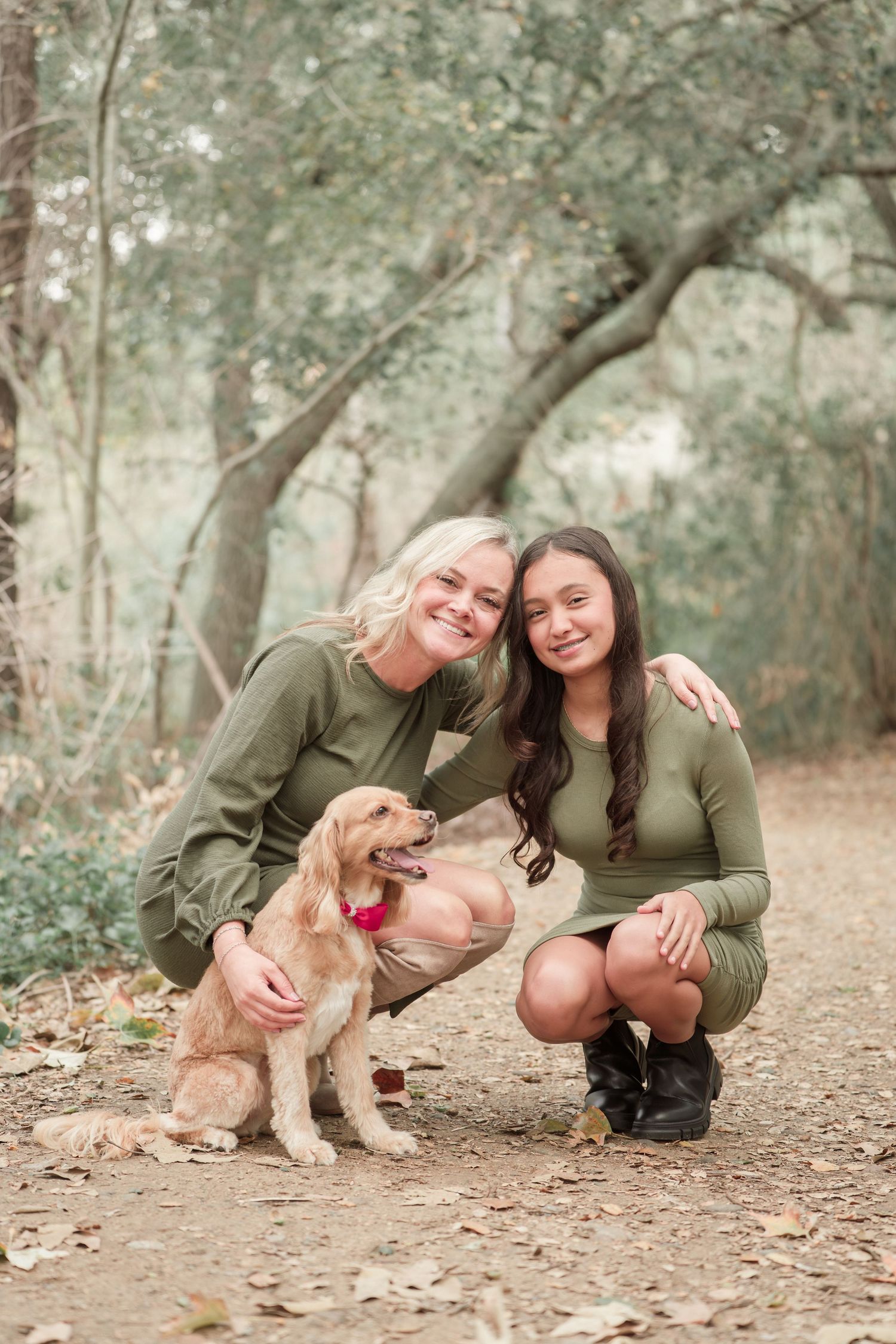 Two women in matching olive green outfits crouch beside a golden Cocker Spaniel in a wooded park setting.