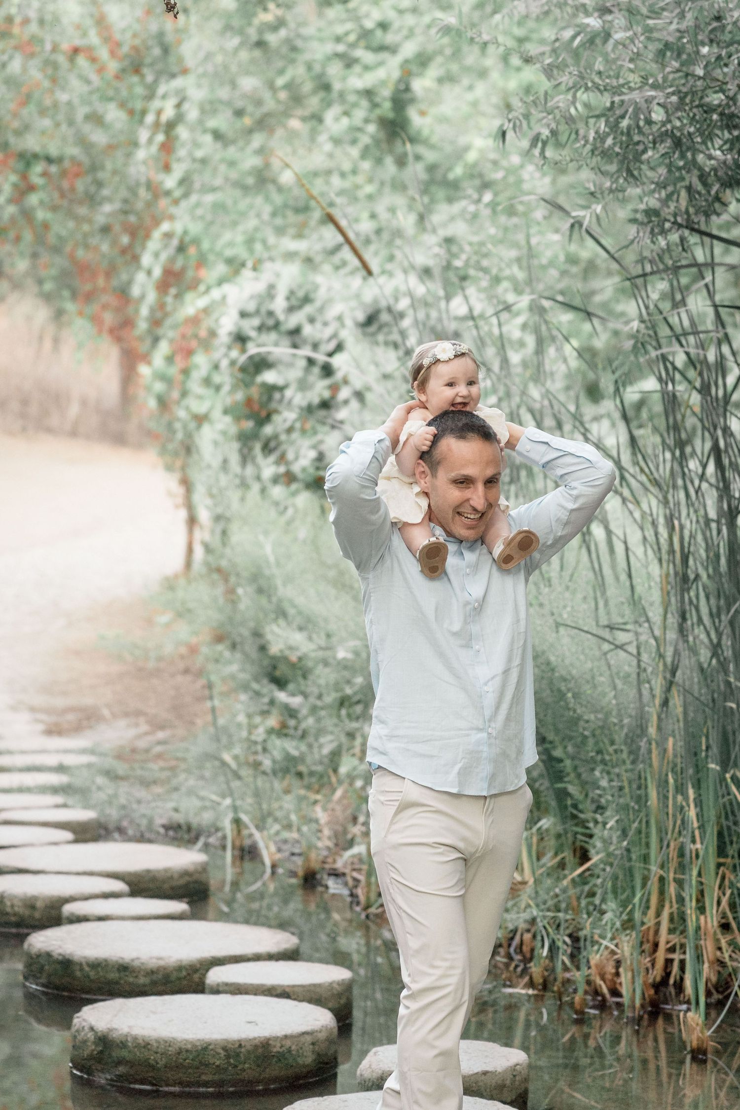 Smiling father carrying baby girl on shoulders while walking on stepping stones in lush garden.